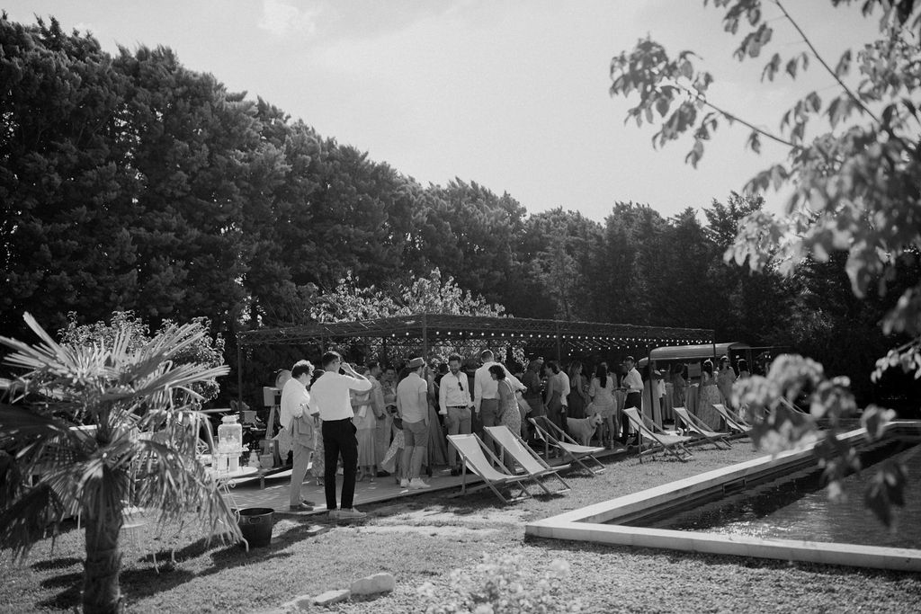 Black-and-white wide shot of guests mingling under fairy-lit pergola beside a swimming pool at cocktail hour