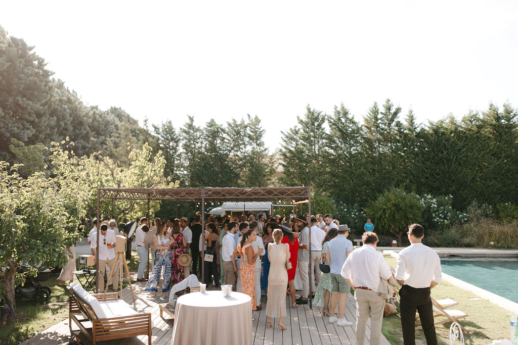 40 guests mingling at poolside cocktail hour with pergola string lights in late afternoon golden light