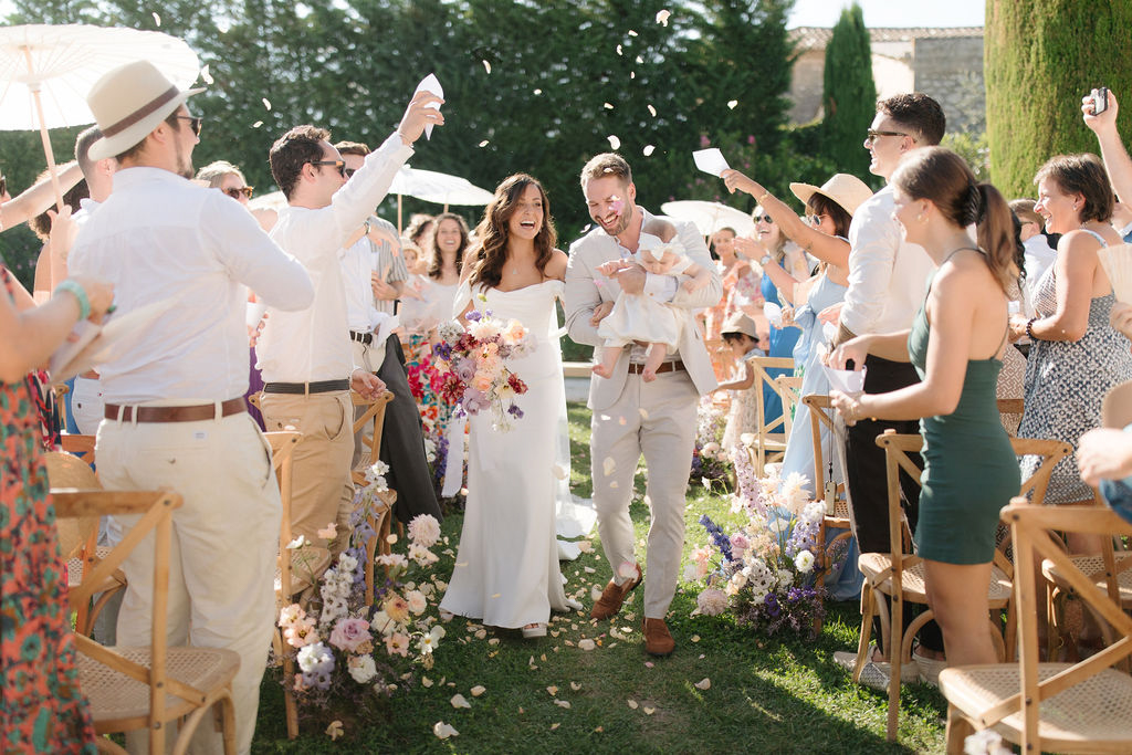 The bride and groom are walking back up the aisle during their outdoor ceremony recessional, surrounded by approximately 40–50 guests who are tossing flower petals and confetti over the couple. The ceremony takes place on a lawn with natural stone architecture visible in the background and tall cypress trees lining the space, suggesting a Provençal or southern French estate. The bride wears a fitted off-the-shoulder white gown and carries a large, loosely arranged bouquet featuring bold pinks, deep burgundy, lavender, and peach blooms including what appear to be dahlias, ranunculus, and sweet peas. The groom is dressed in a light sand-colored suit with brown leather shoes and holds a small child. Aisle arrangements of white, blush, lavender, and peach florals are placed along the ground, with loose petals scattered across the grass. Several guests hold white parasols to shade from the bright sun, and seating consists of natural wood cross-back chairs. The guests' attire is varied and colorful, including a dark green dress and floral prints. This is a wide mid-shot taken at aisle level, capturing the couple centered with motion and energy from the petal toss.