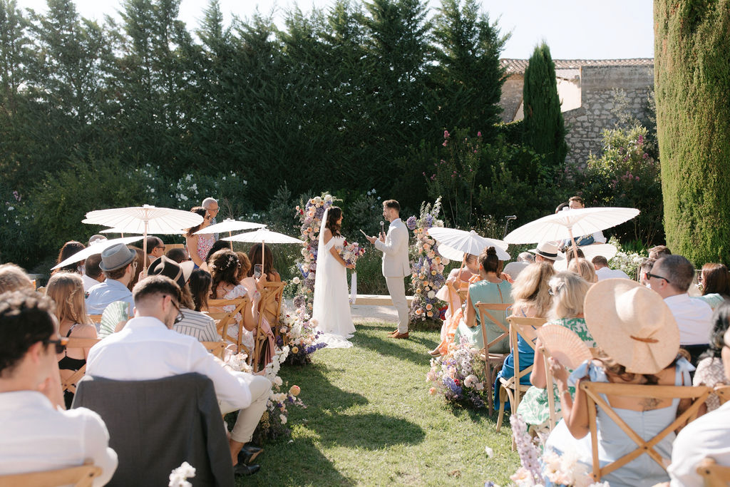 An outdoor wedding ceremony taking place in a walled garden, likely in the south of France, with a stone building visible in the background. The bride wears a white off-shoulder dress with a veil, and the groom is dressed in a light beige suit; they stand at an altar framed by two tall floral columns featuring a colorful mix of peach, yellow, purple, and white blooms including what appear to be ranunculus, delphinium, and wildflower varieties, with matching florals lining the aisle. Approximately 40–50 guests are seated on wooden folding chairs on either side of a grass aisle, many holding white paper parasols and hand fans to shade themselves from the bright sun, dressed in summery, colorful attire. The overall styling is relaxed and garden-party in feel, with a soft, wildflower-inspired floral palette; the shot is taken from the back of the aisle as a wide perspective view looking toward the couple and officiant.