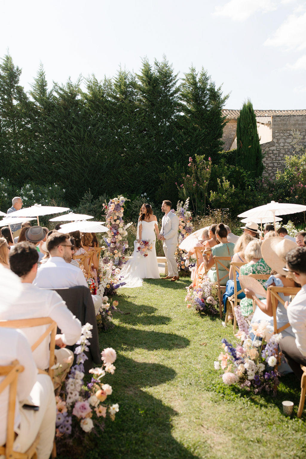 Outdoor garden ceremony with colorful floral aisle and guests holding parasols in Provencal setting