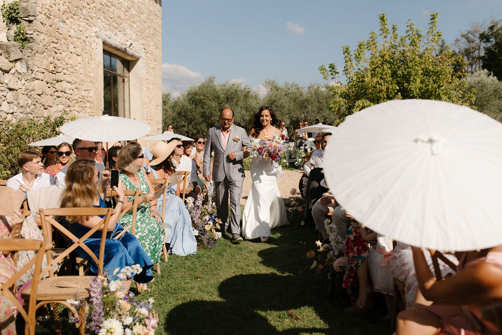 An outdoor wedding ceremony processional captured in a wide shot, showing a bride walking down the aisle escorted by a man in a light grey suit with a pink shirt, likely her father. The bride wears a fitted white sleeveless gown and carries a large, loosely arranged bouquet of mixed wildflower-style blooms in purples, pinks, yellows, and whites. The ceremony takes place on a grass lawn alongside a historic stone building, with wooden cross-back chairs lining the aisle for approximately 30–40 seated guests. Several guests are holding white paper parasols to shade themselves from the bright sun, and many wear colorful summer attire including floral prints, blue, and green dresses. Loose floral arrangements in purples, whites, and soft pinks are placed along the aisle edge, complementing the wildflower bouquet. The overall styling is relaxed and summery with a garden-party feel.
