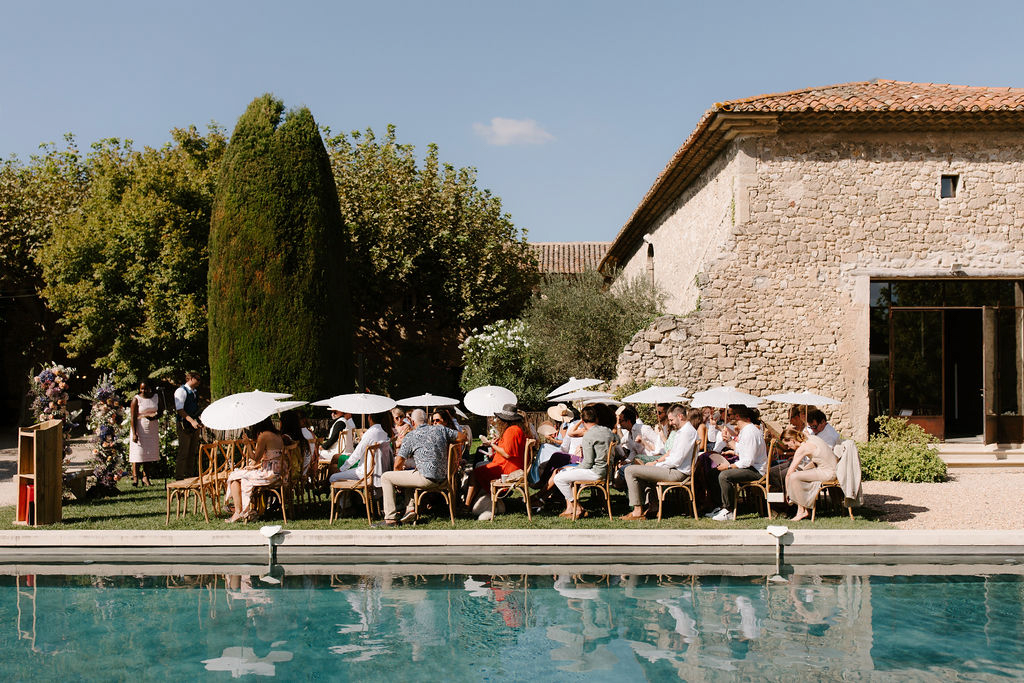 An outdoor wedding ceremony taking place alongside a rectangular swimming pool at a French stone property with terracotta roof tiles. Approximately 25–30 guests are seated in rattan bistro chairs arranged in rows along the poolside, with several white parasols open above them to provide shade in the bright afternoon sun. Guests are dressed in summer attire in a mix of neutral tones, white, and a few pops of orange and coral. To the left of the seating, a floral installation in muted tones of dried and fresh flowers in purples, blues, and pinks is partially visible, along with what appears to be an officiant or couple standing near a wooden lectern. The wide shot captures the full scene reflected in the pool water in the foreground, with the venue's aged limestone building and formal garden with a tall cypress tree visible in the background. Potential venue feature image.