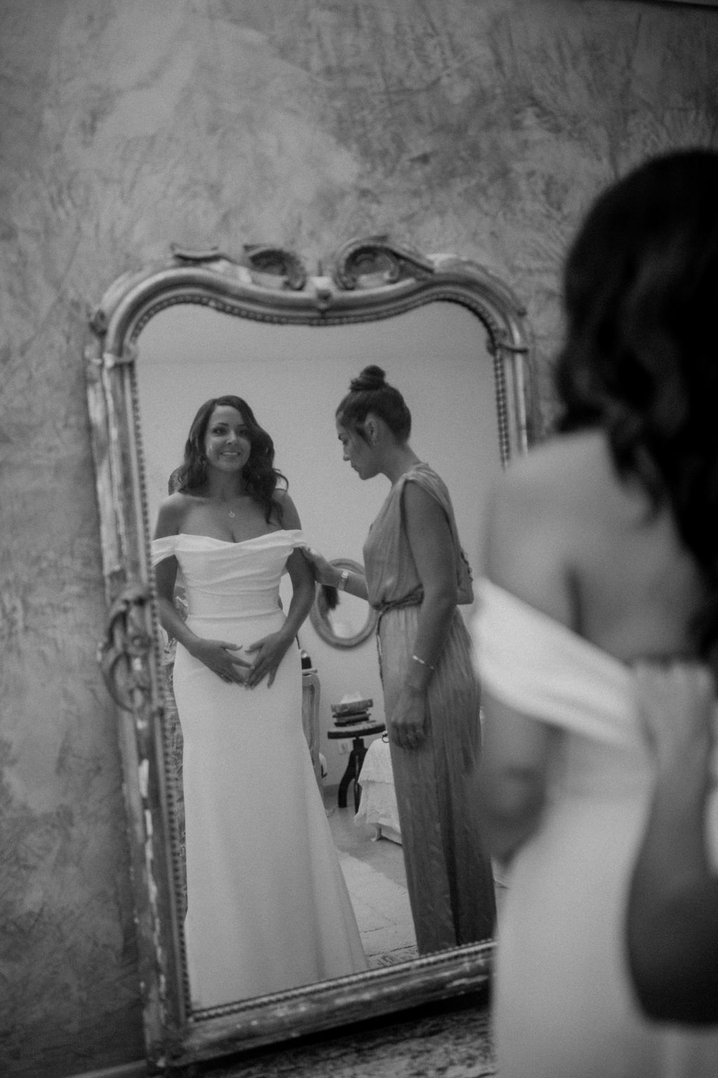 Black and white bride smiling at mirror reflection as attendant adjusts minimalist off-shoulder gown