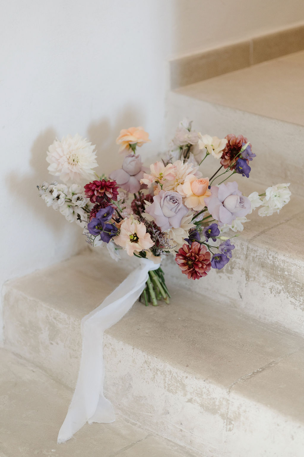 Bridal bouquet of lavender roses, peach ranunculus, and burgundy dahlias with trailing white ribbon on stone steps