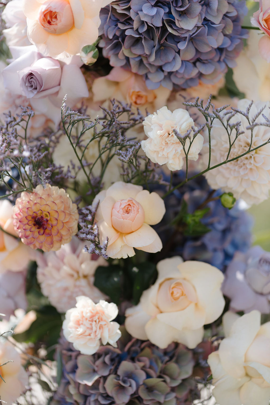 Dense floral arrangement of dusty blue hydrangeas, blush garden roses, peach dahlias, and mauve roses
