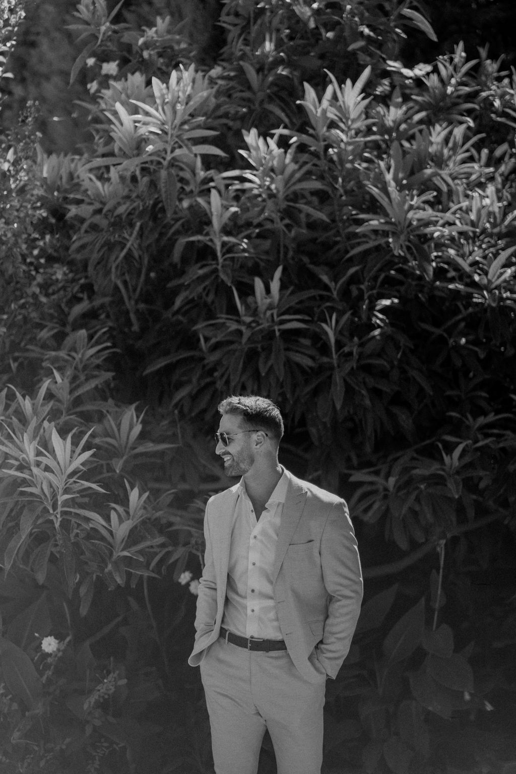 Black and white portrait of groom in light suit and aviator sunglasses with hands in pockets in garden