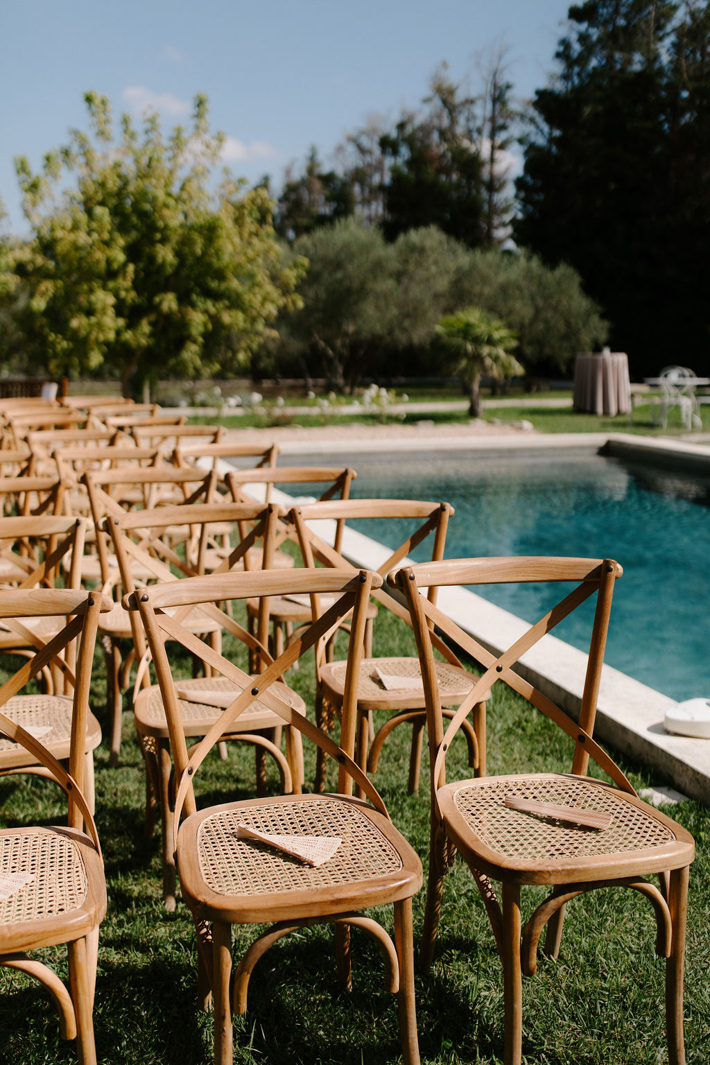 Outdoor ceremony setup with cross-back chairs holding blush paper fans arranged beside a swimming pool on a lawn