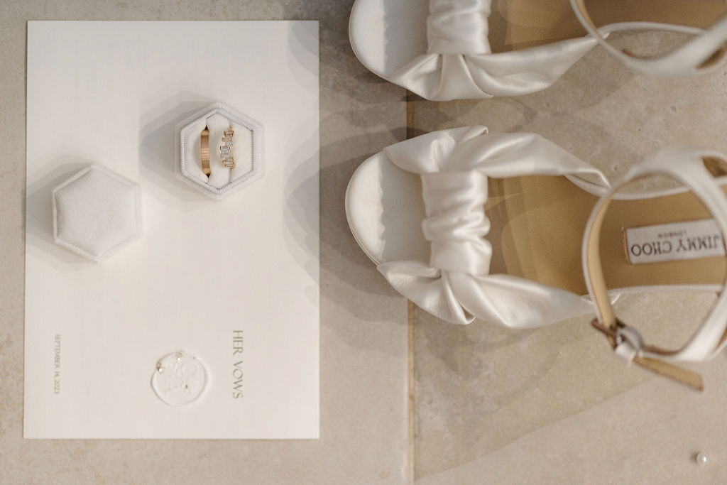 Flat lay of vow booklet hexagonal ring box with gold bands and white Jimmy Choo heels