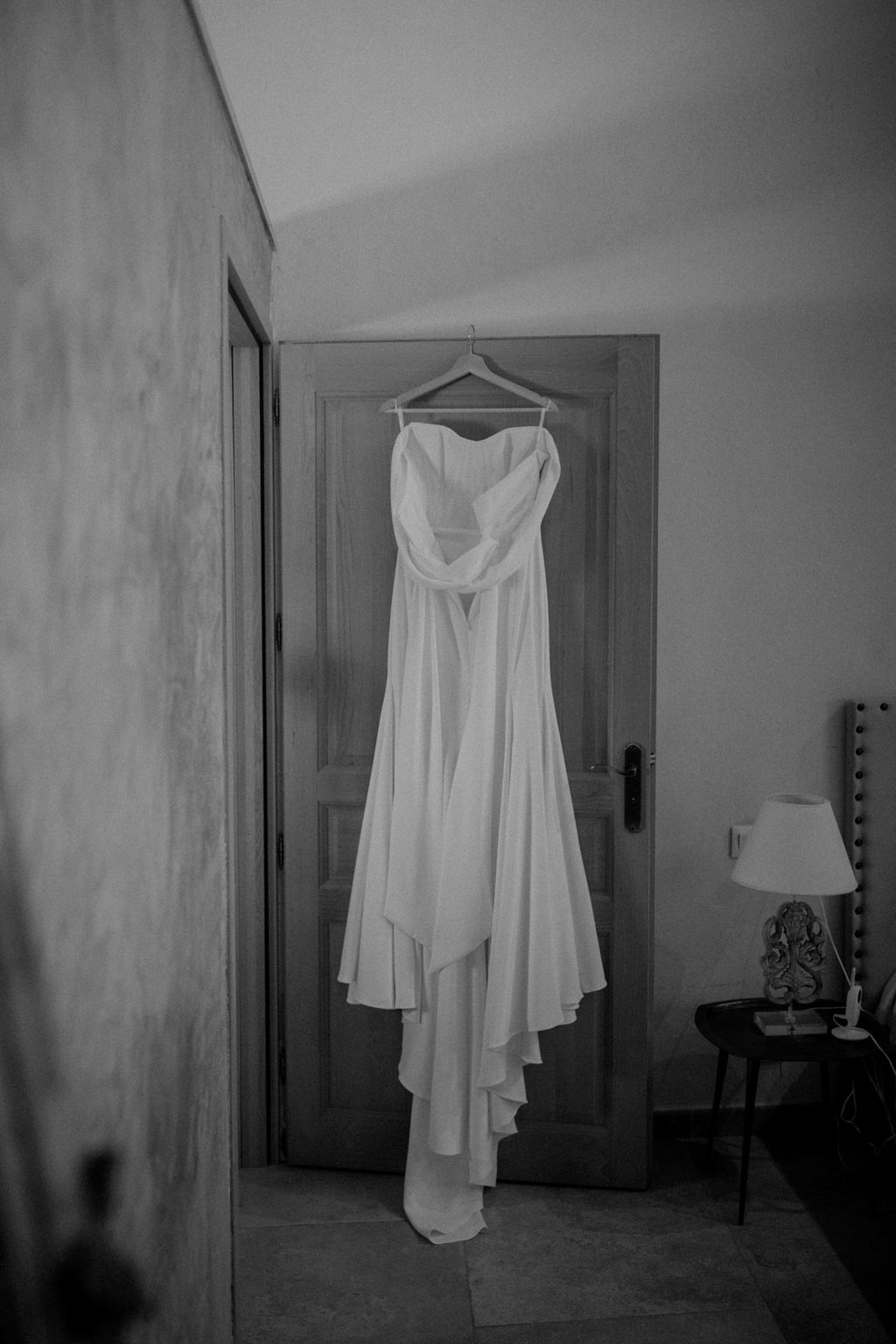 This black-and-white getting-ready detail shot shows a strapless wedding dress hanging from a wooden hanger on a door frame in what appears to be a château or period property room. The gown features a draped sweetheart bodice with flowing, layered skirt panels that trail toward the floor, suggesting a light, fluid fabric such as silk or chiffon. The room interior shows a vaulted ceiling, aged plaster walls, a small bedside table with a table lamp, and a vanity mirror with bulb lighting partially visible on the right. The image has soft contrast with bright mid-tones on the dress against the darker door and room surroundings.