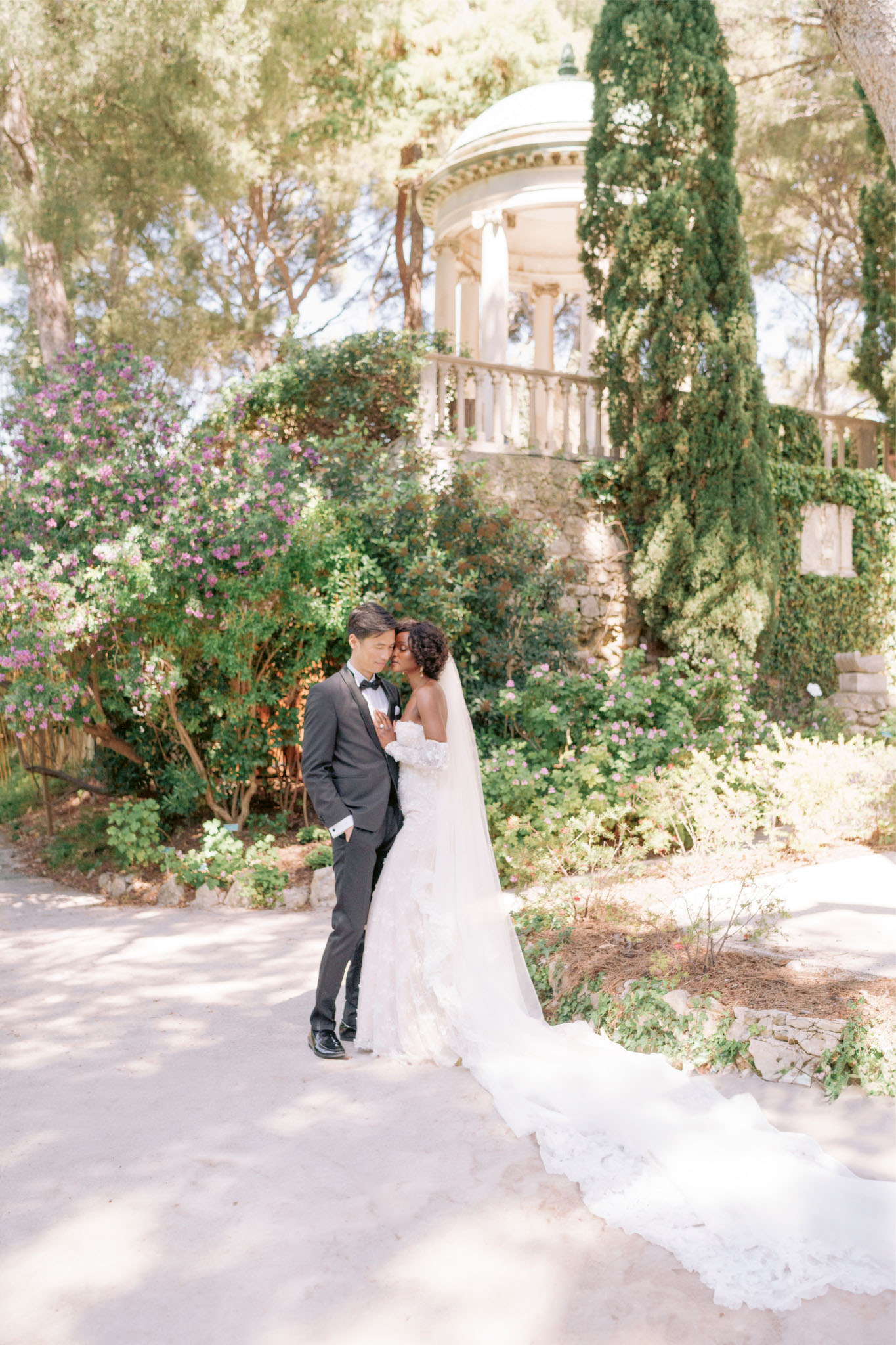 Bride in off-the-shoulder lace ball gown and groom in charcoal tuxedo touching foreheads before stone rotunda