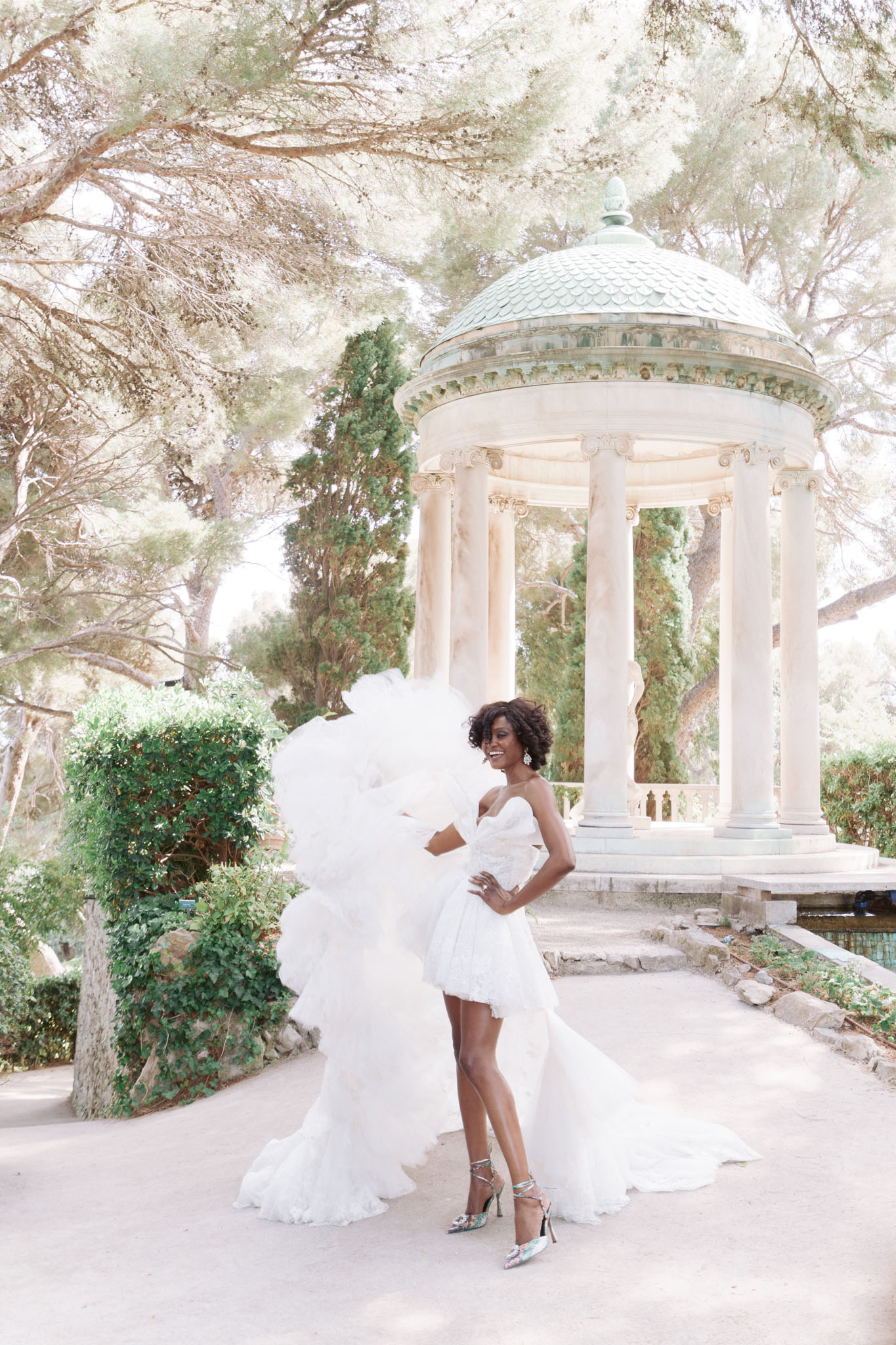 A bridal portrait taken outdoors in the grounds of a formal garden, featuring a bride posing solo in front of a classical white marble rotunda with Ionic columns and a verdigris copper domed roof. The bride wears a white strapless sweetheart mini wedding dress with voluminous ruffled tulle detailing at the shoulders and sides, paired with metallic silver strappy heeled sandals and statement earrings; the dress also has a floor-length train pooling behind her. She stands in a confident pose with one hand on her hip and is smiling. The image is a full-length portrait shot with soft, bright natural lighting giving the overall image a pale, airy tone. Potential venue feature image.