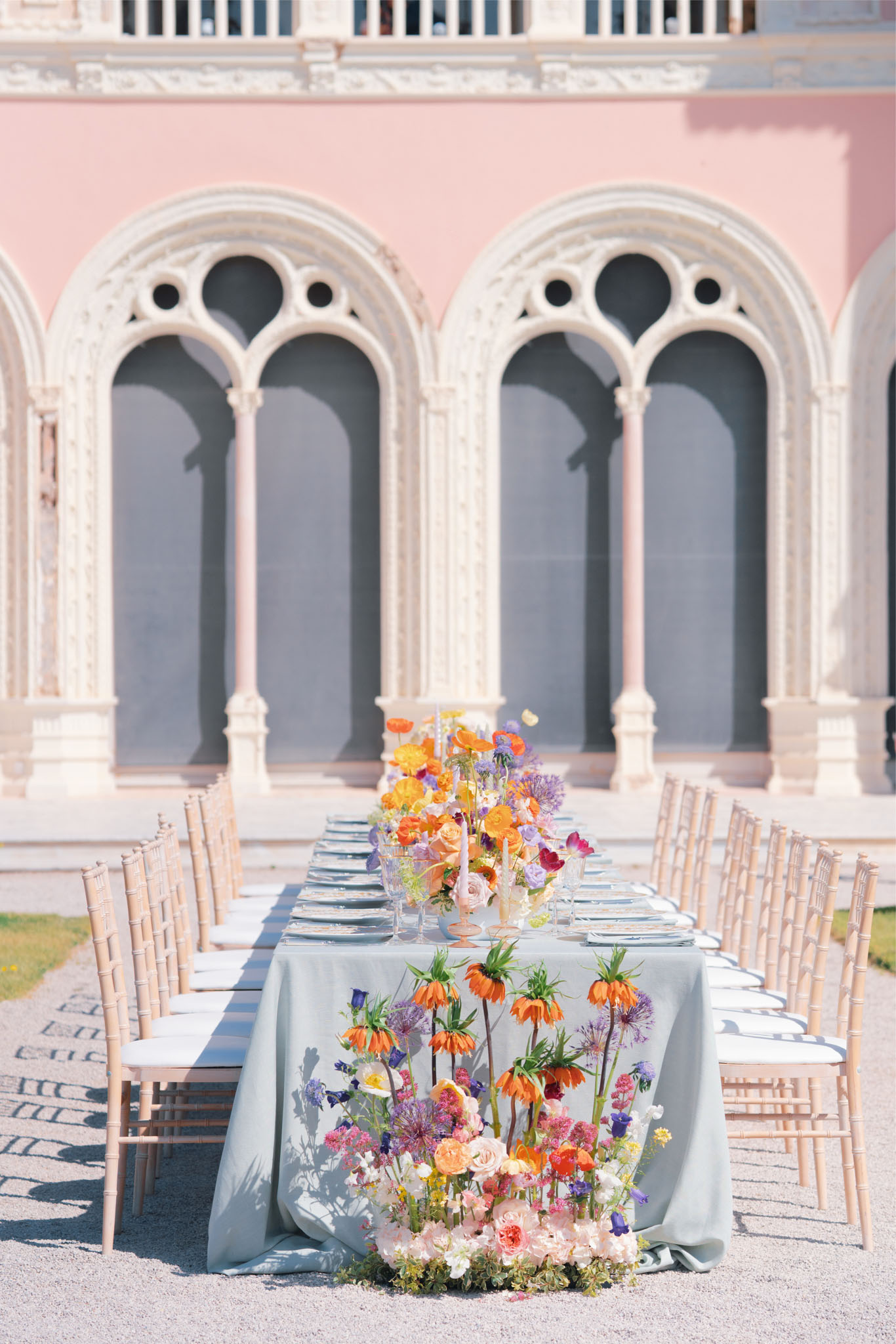 An outdoor reception tablescape set in front of a pink-facades building with ornate white Gothic-style arched windows and colonnades. The long rectangular dining table is dressed in a dusty sage blue linen and flanked by natural wood chiavari chairs with white seat pads. The table is set with gray charger plates and glassware running the full length. A lush floral runner cascades across the table and spills down onto the ground at the near end, featuring orange crown imperial fritillaria, blush garden roses, purple allium, lavender scabiosa, orange ranunculus, hot pink sweet peas, and yellow blooms arranged in a loose, garden-style composition. A second floral arrangement is visible mid-table, continuing the same vibrant multicolor palette of orange, purple, pink, and yellow. The overall styling is colorful and maximalist with a Mediterranean-influenced backdrop. Wide portrait-orientation shot. Potential venue feature image.