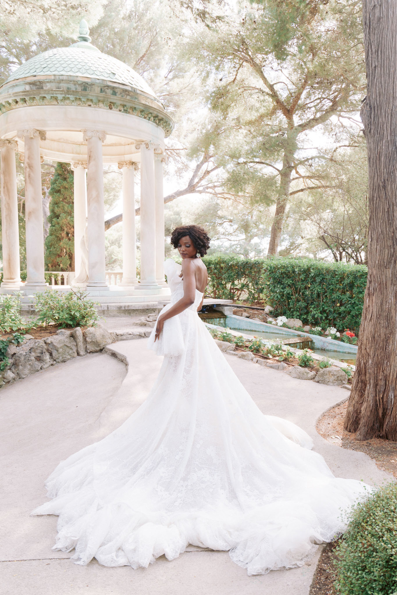 A bridal portrait taken outdoors in a formal garden setting, featuring a single bride posed on a curved stone pathway. She wears a strapless white ball gown with a heavily layered lace and tulle skirt and an exceptionally long, full cathedral-length train that spreads across the path behind her. She is turned slightly away from the camera, glancing back over her shoulder, with natural curly hair and statement earrings. In the background stands a circular neoclassical rotunda pavilion with white Corinthian columns and a verdigris copper dome roof, alongside a narrow rectangular ornamental pool edged with flowering plants. The overall styling is classic and formal. The image is a full-length portrait shot with soft, airy natural light. Potential venue feature image.