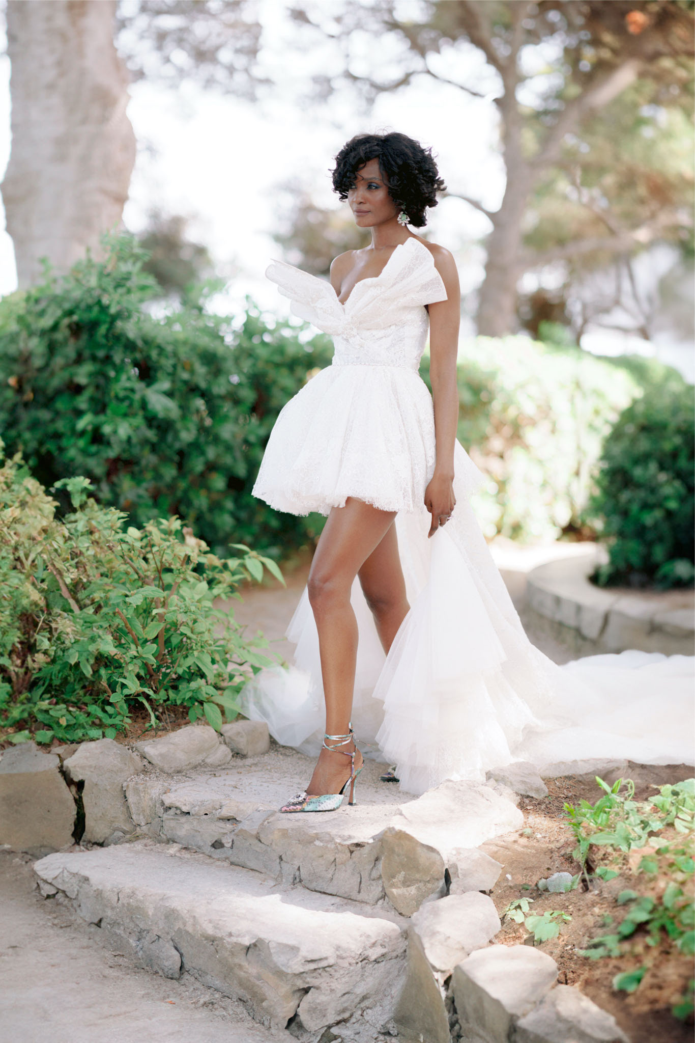 A bridal portrait shot outdoors in a garden setting, featuring a single bride standing on rustic stone steps surrounded by greenery. She wears a white structured mini dress with a full textured skirt, a dramatic oversized bow detail at the neckline, and a long tulle train flowing behind her. Her accessories include statement drop earrings and embellished silver strappy heeled pumps with a pointed toe. Her hair is short and curly, worn down. The composition is a full-length portrait with a shallow depth of field, softly blurring the garden background. The overall styling aesthetic is modern and fashion-forward.