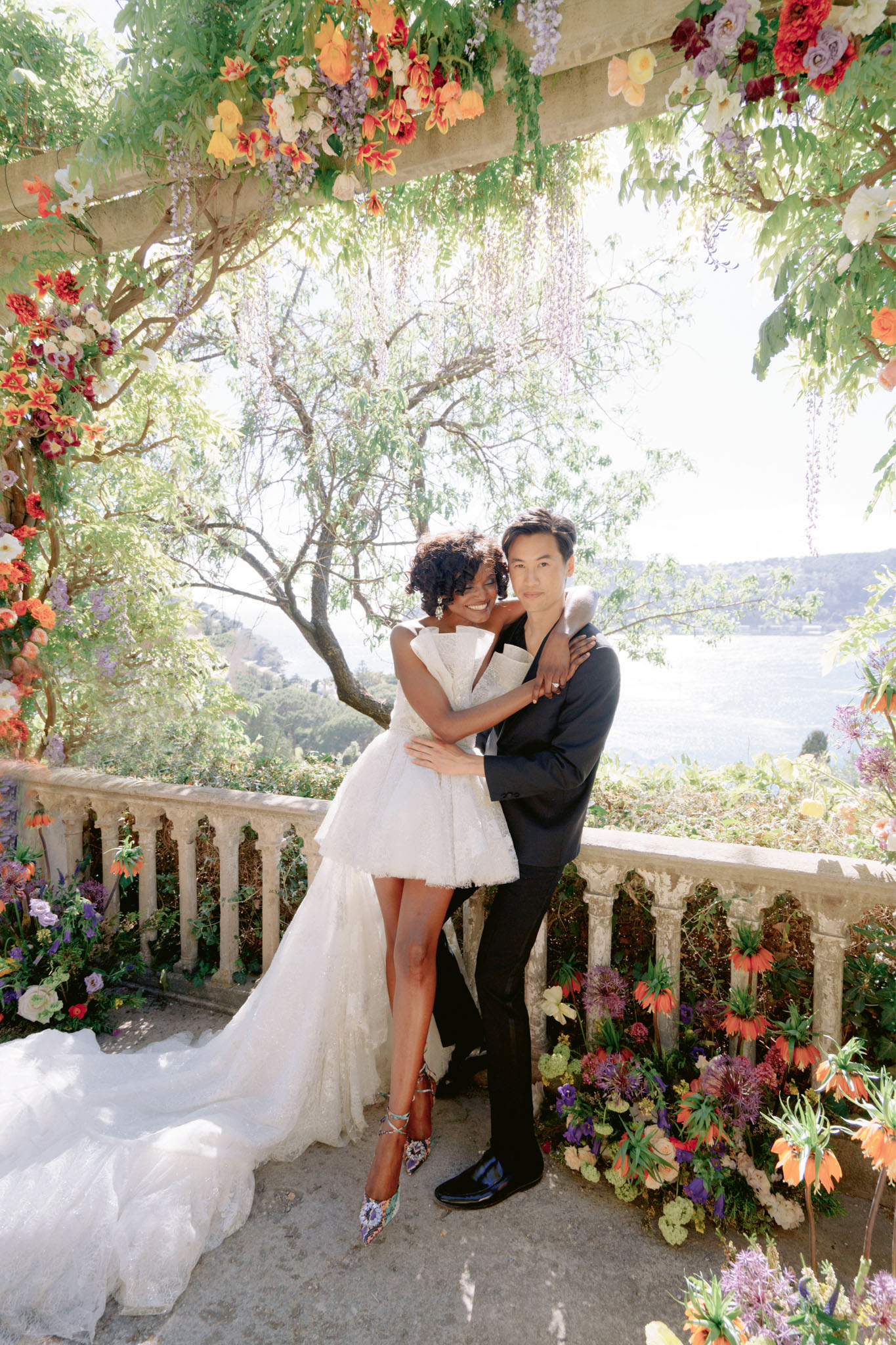 Bride in white strapless mini dress and groom in black suit posing under cascading floral arch on stone terrace