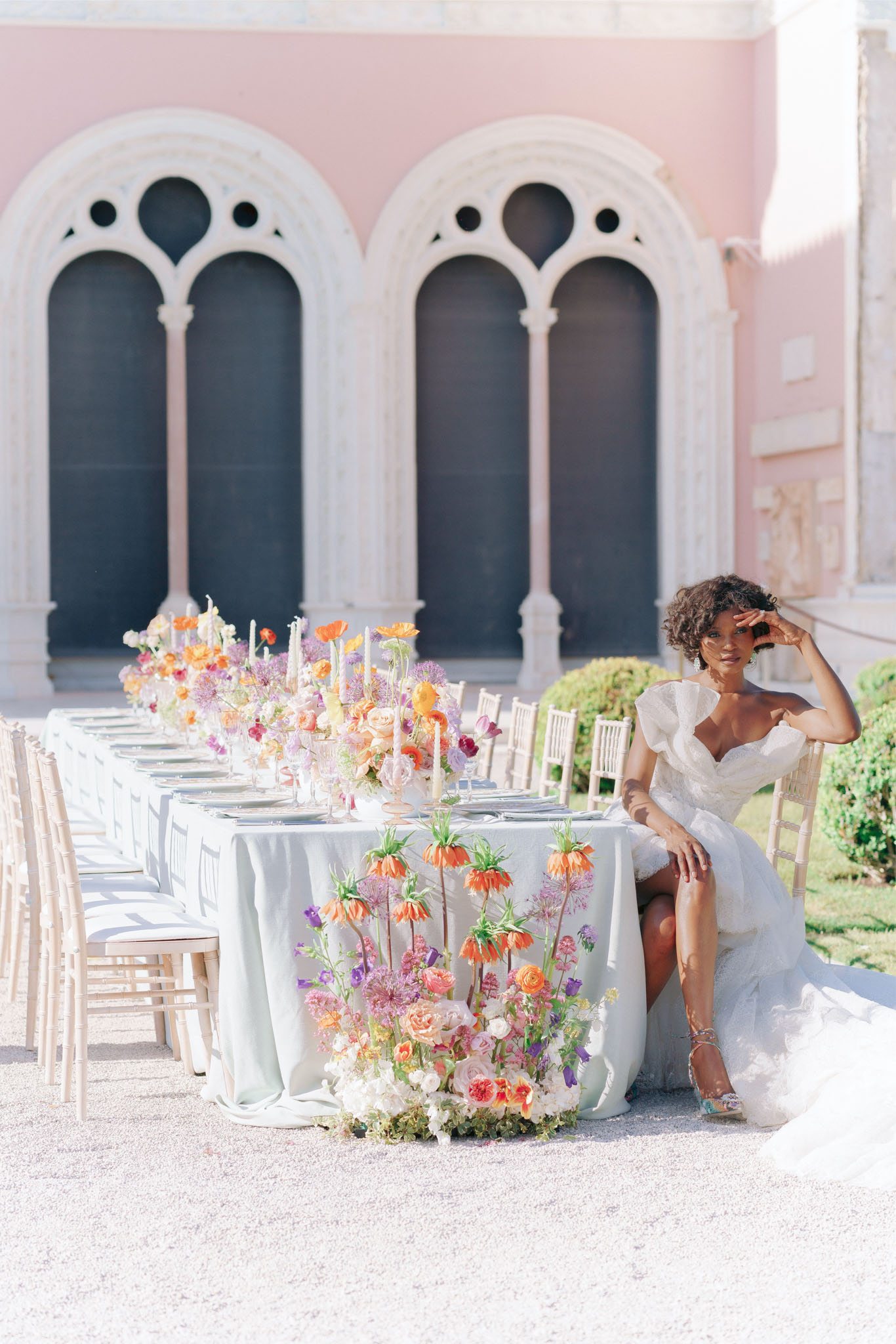 Bride at sage-linen table with orange poppy and coral rose runner beside pink Gothic villa