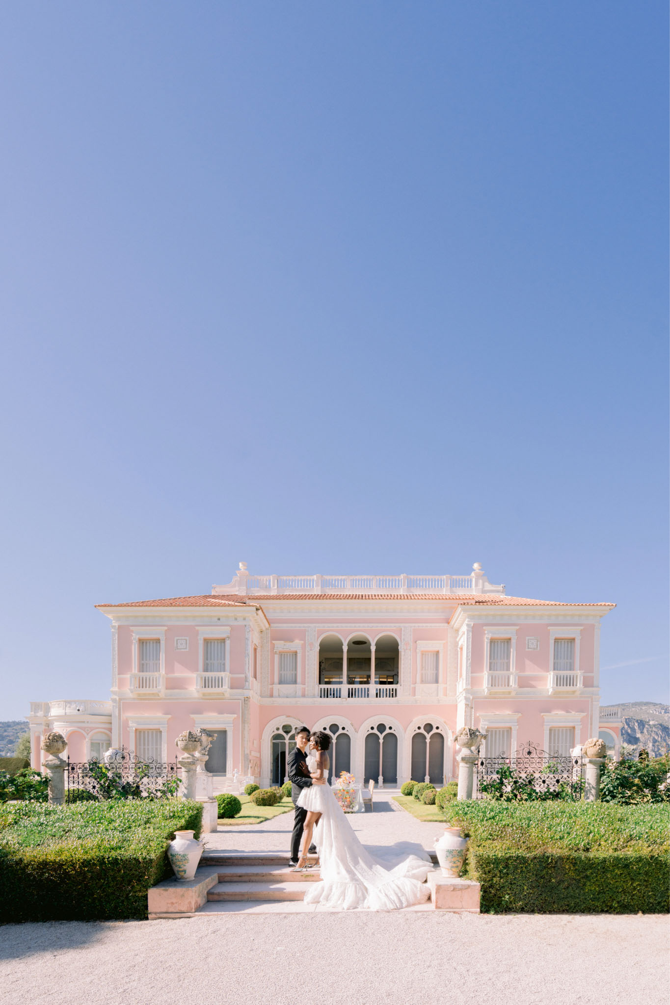 Bride and groom embracing on steps of pink Belle Epoque villa with boxwood hedges and topiary