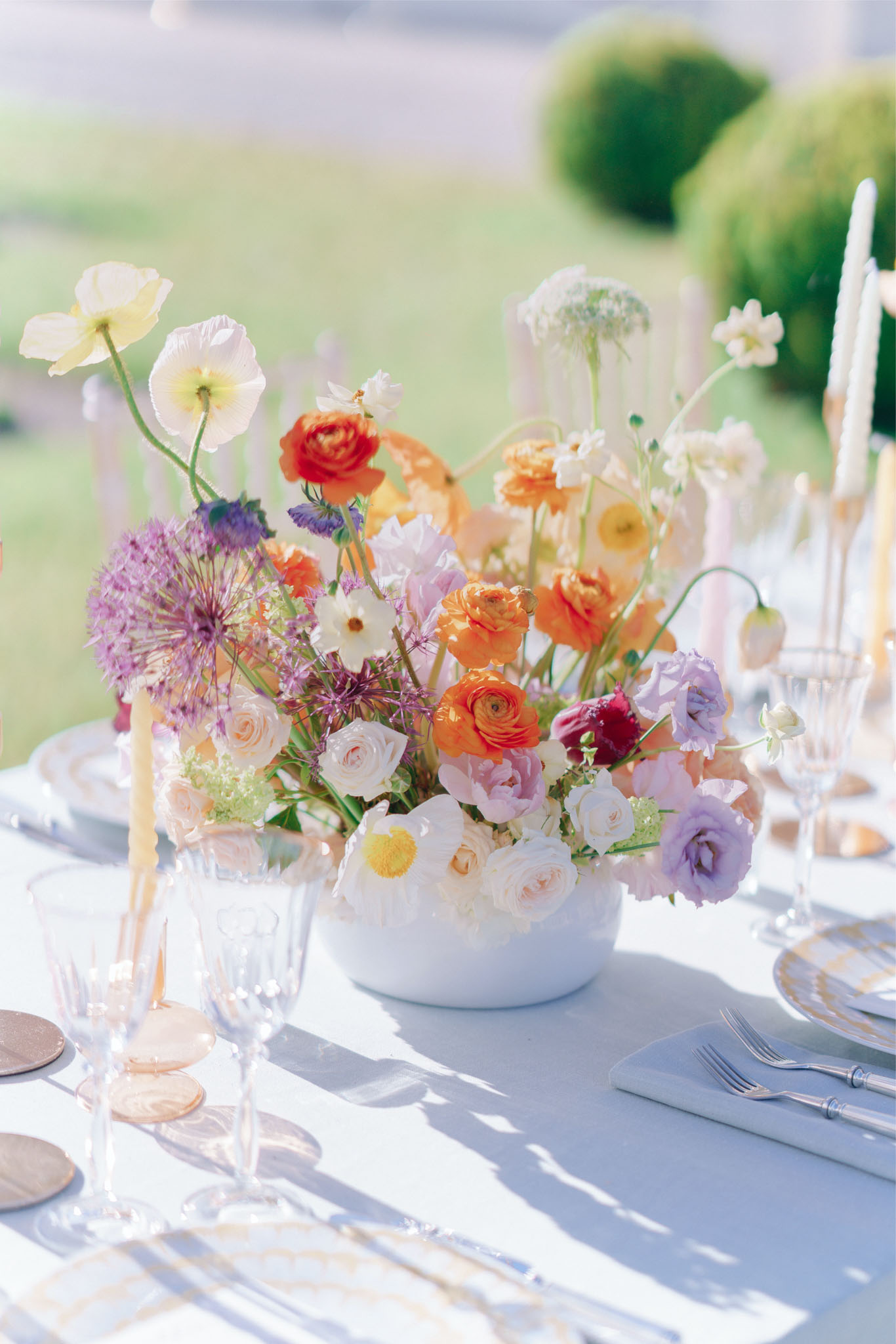 Wildflower centerpiece of orange ranunculus purple allium yellow poppies in white bowl on gold chargers
