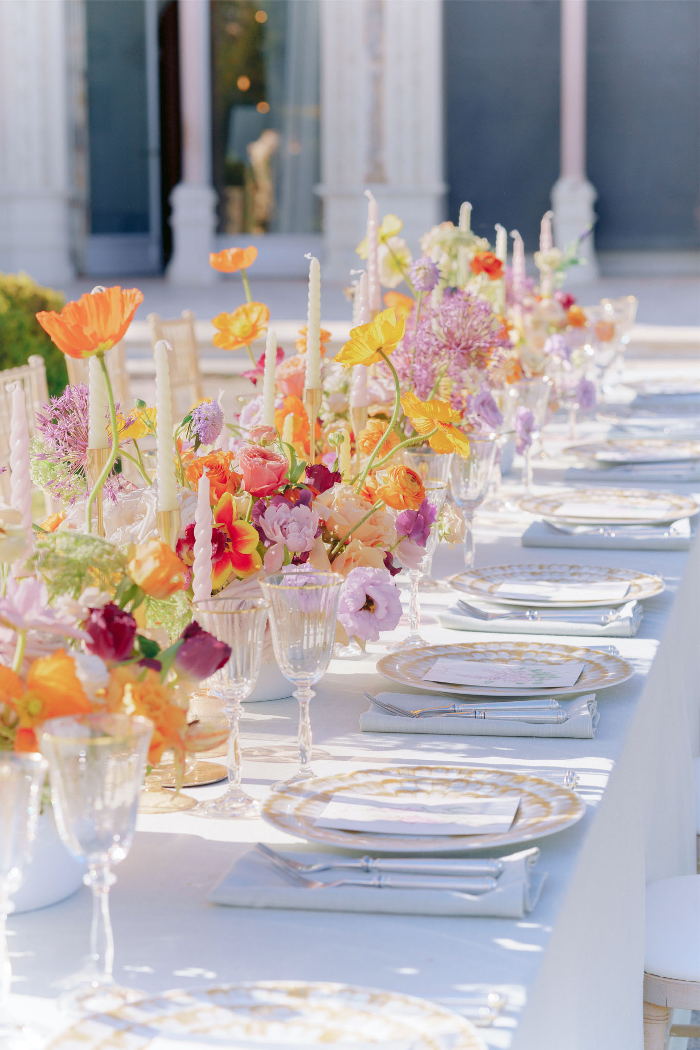 A close-up detail shot of an outdoor wedding reception table set against what appears to be a classical building with white columns visible in the background. The long table is dressed in a white linen tablecloth and set with stacked place settings featuring white plates with gold-trimmed borders, folded white linen napkins, silver cutlery, printed menu cards, and clear crystal glassware with twisted stems. The floral centerpieces are abundant and run the length of the table, featuring orange Iceland poppies, yellow ranunculus, coral and red roses, lavender lisianthus, purple allium, and mixed blooms in a vivid multicolor palette of orange, yellow, coral, magenta, lavender, and cream. Tall ivory and pale pink twisted taper candles are interspersed throughout the florals. The overall styling is bright and colorful with a garden-party aesthetic, and gold chiavari chairs are partially visible along the side of the table.
