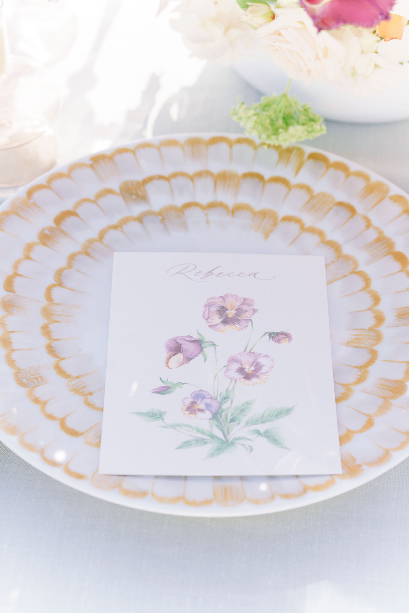 Close-up detail shot of a wedding reception place setting. A white charger plate with a gold scalloped border holds a square place card reading 'Rebecca' in script calligraphy, illustrated with a watercolor botanical motif of purple and lavender pansies with green foliage. The table is dressed with a light grey or white linen, and a floral centerpiece featuring blush garden roses, white blooms, and green accents is partially visible in the background. The overall decor palette combines gold, white, and soft lavender with a botanical, garden-inspired styling theme.