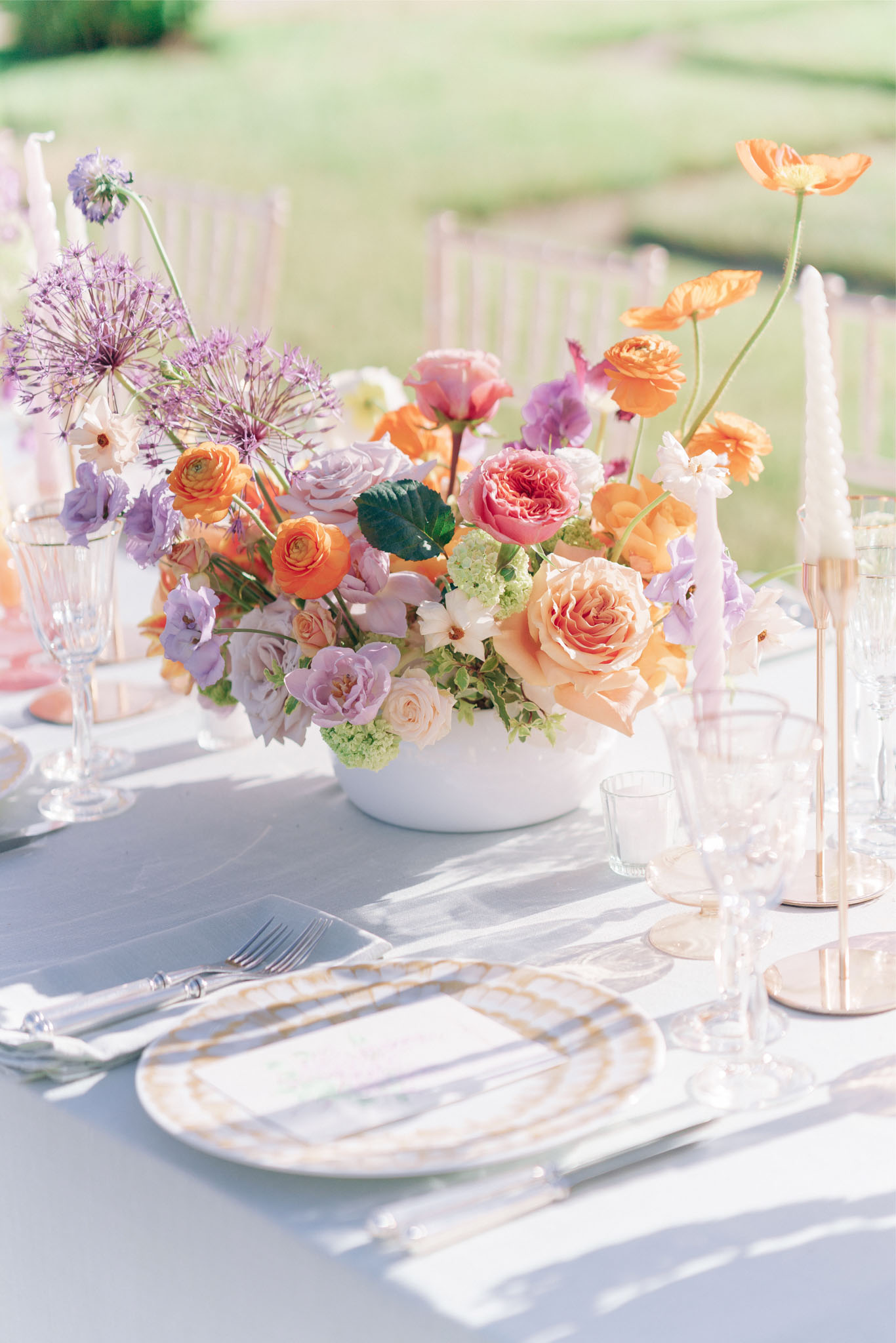 Outdoor reception table with orange and lavender floral centrepiece, gold charger plates, and taper candles