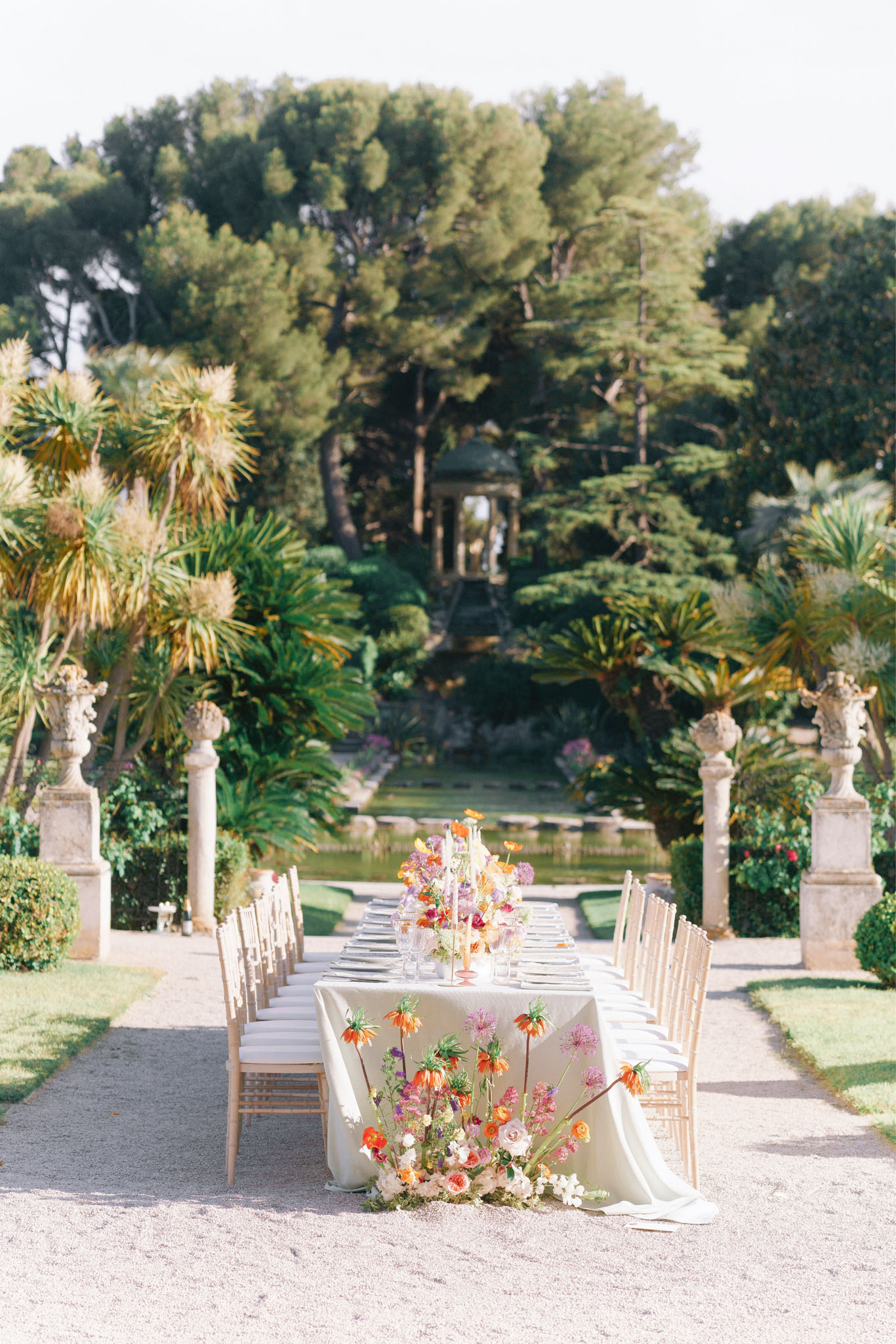 An outdoor reception table setup photographed in a wide shot along a gravel garden path leading toward a formal garden with a reflecting pool and a stone gazebo in the background. The long rectangular table is dressed in an ivory linen tablecloth that drapes to the ground, set with place settings and flanked by gold Chiavari chairs on both sides, seating approximately 16–18 guests. A large, loose floral arrangement cascades from the near end of the table onto the ground, featuring orange crown imperial fritillaria, pink alliums, peach and ivory ranunculus, purple sweet peas, and white blooms, with tall taper candles in amber-toned candlestick holders at the center of the table. Stone pedestals topped with ornamental urns frame the path on either side, and the formal garden design with trimmed hedges, palm-like plants, and mature trees contributes to a classic, structured French estate aesthetic. Potential venue feature image.