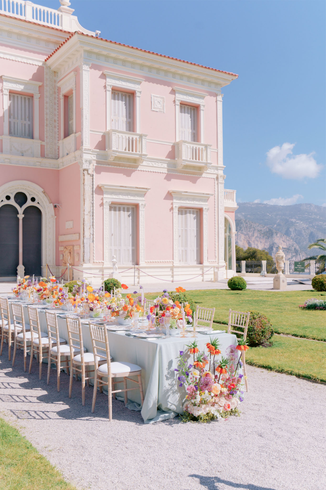 Long reception table with sage green linen and colorful floral runner set on terrace before pink Belle Epoque villa