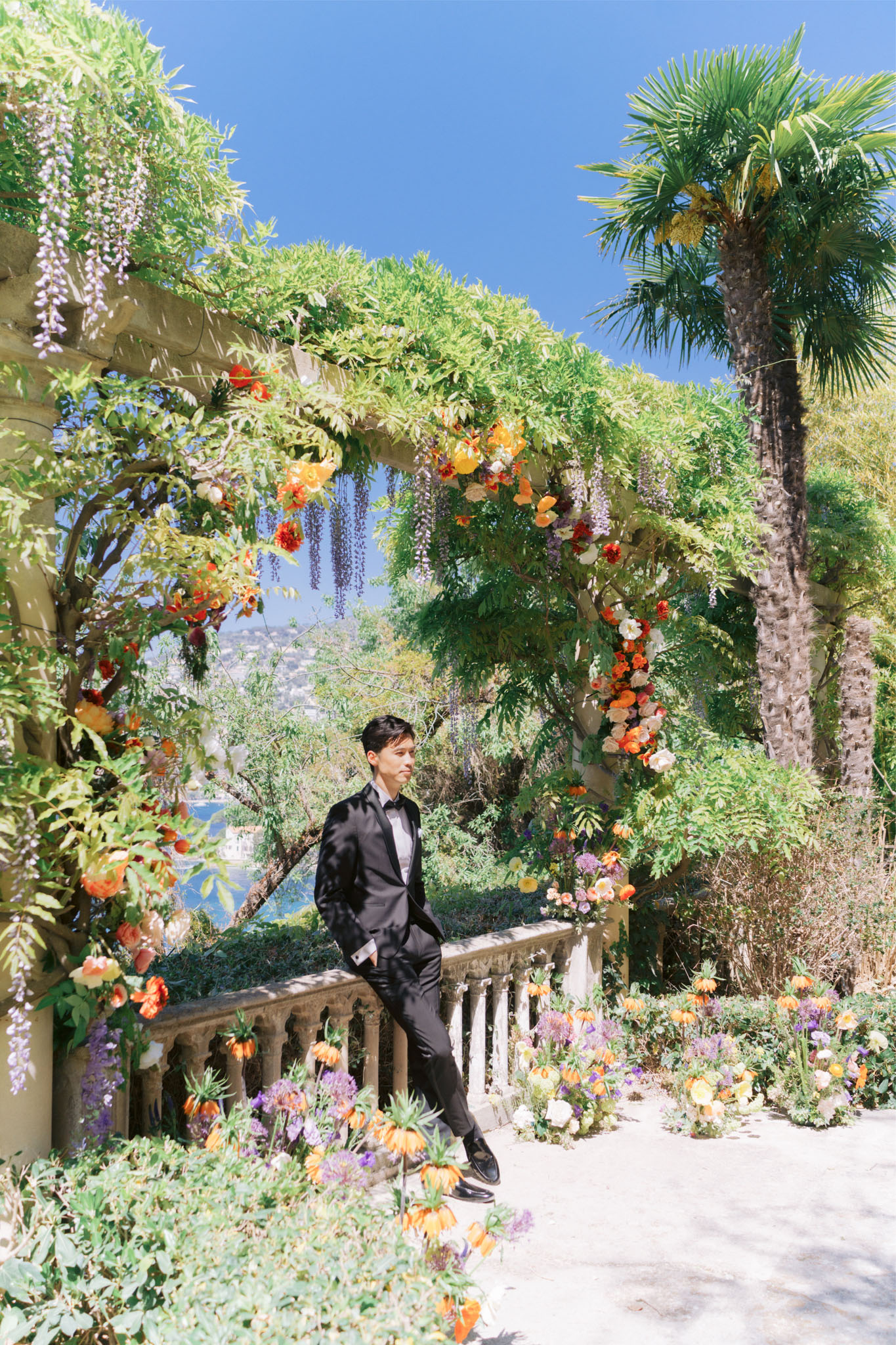 Groom in black tuxedo standing under wisteria-draped pergola with orange and red floral arrangements