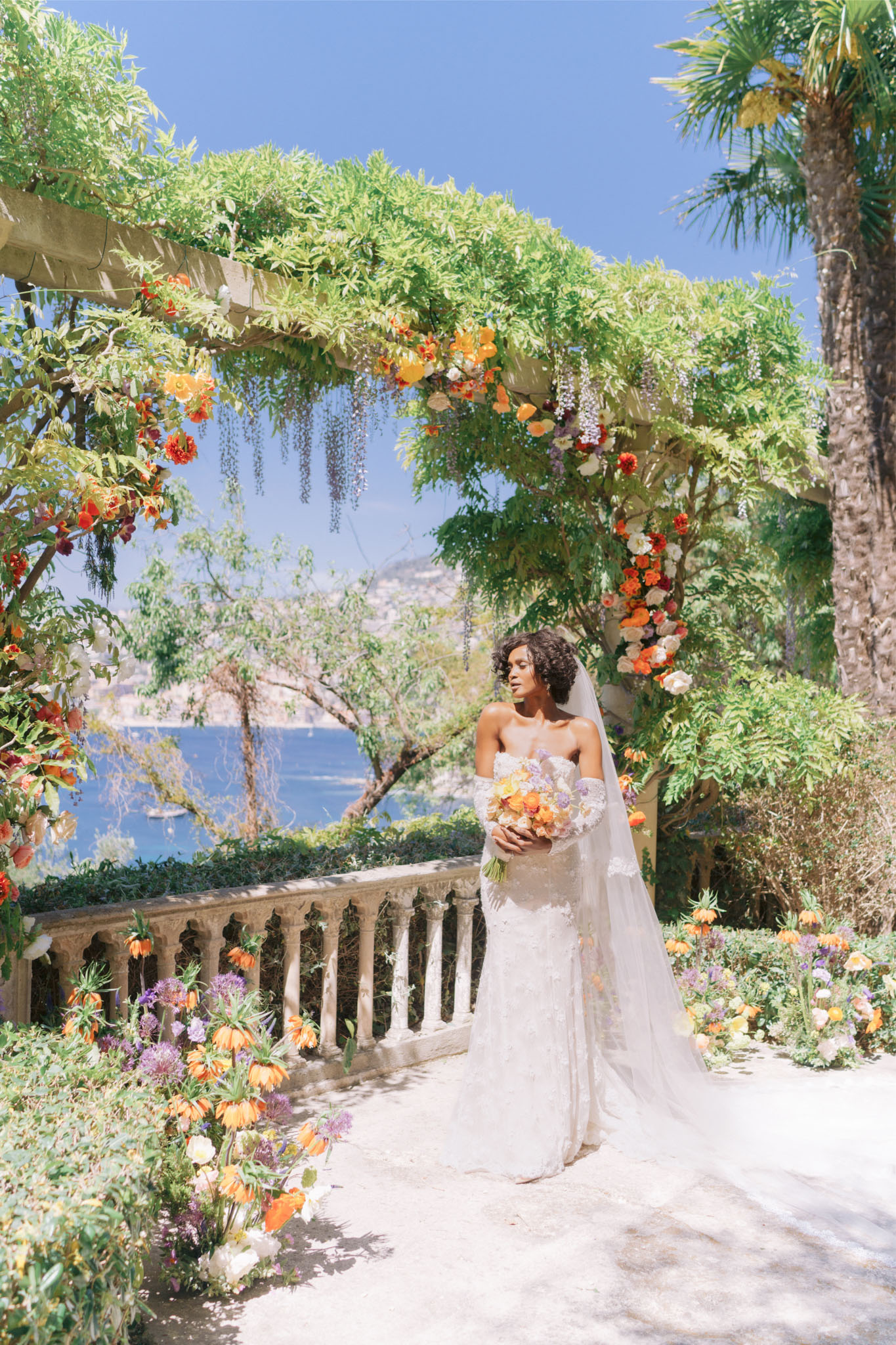 Bride under vine pergola with vibrant orange, red, and lavender floral arch overlooking Mediterranean lake