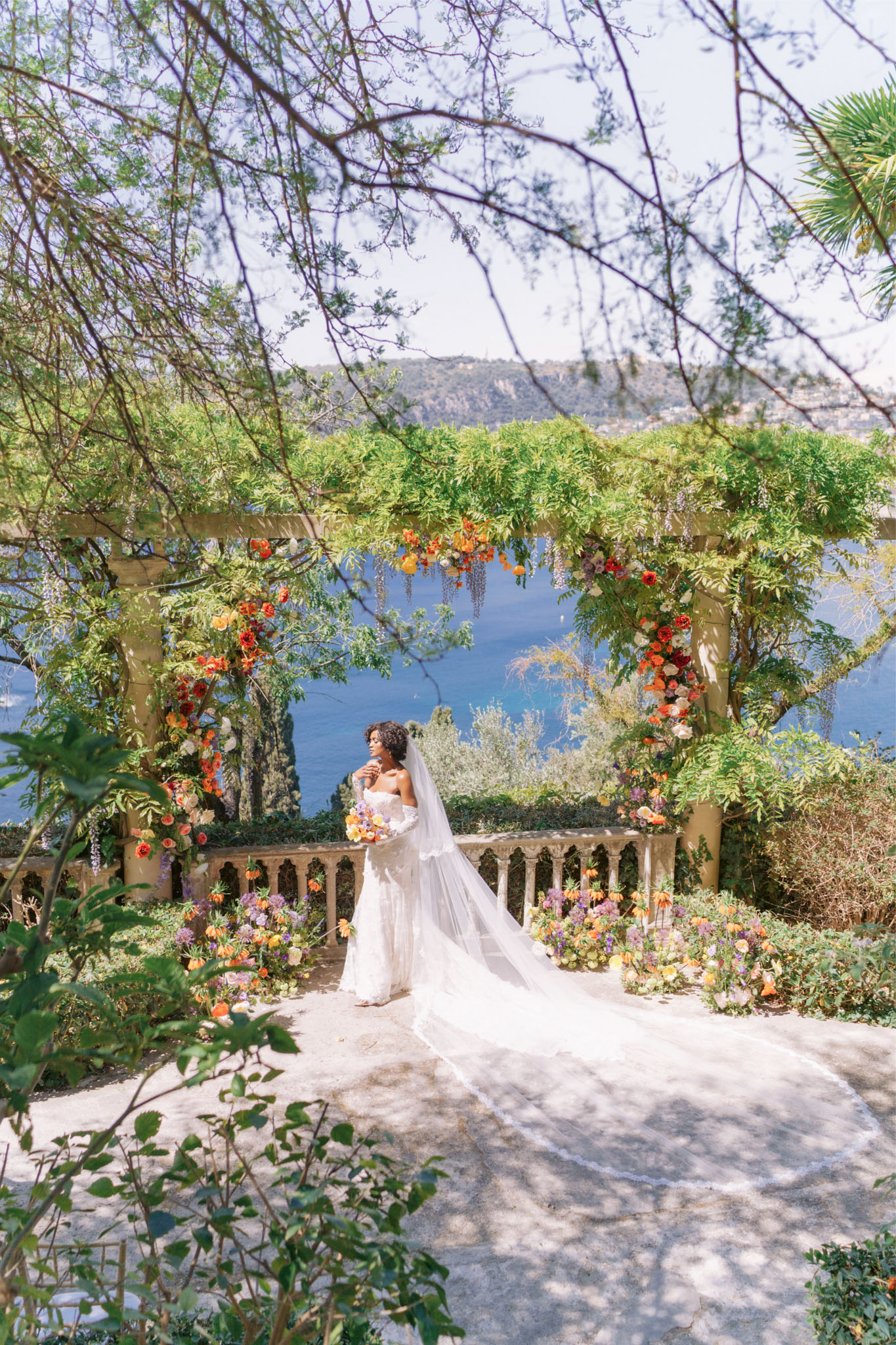 Bride under vine pergola with orange and purple floral arch holding colourful bouquet overlooking coast
