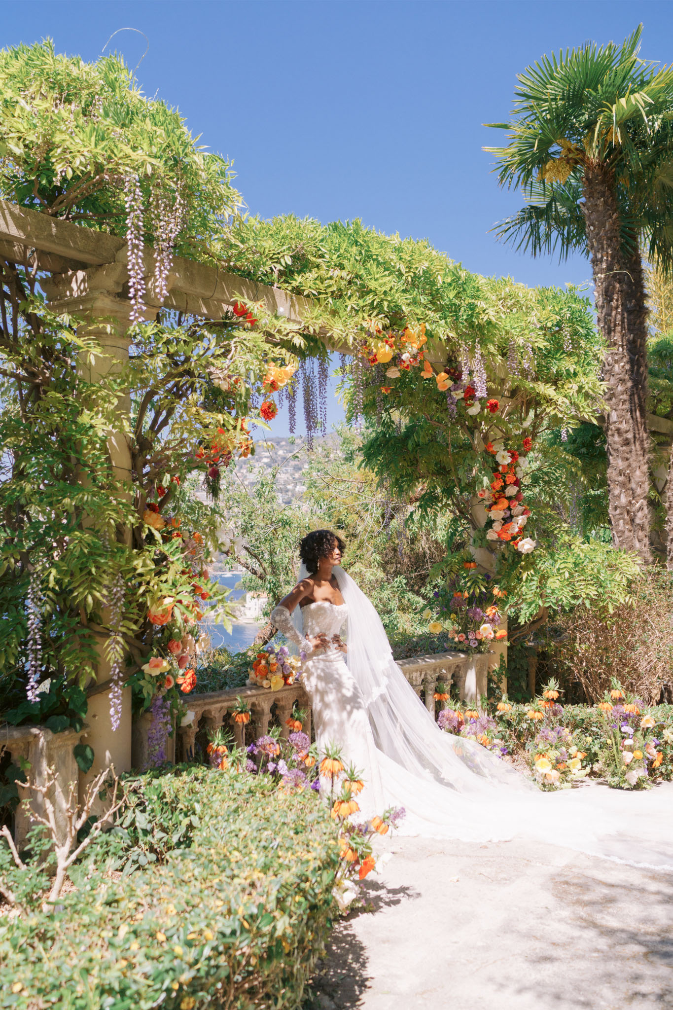 Bride on vine-covered terrace with orange ranunculus and lavender wisteria floral arch overlooking coastal hillside