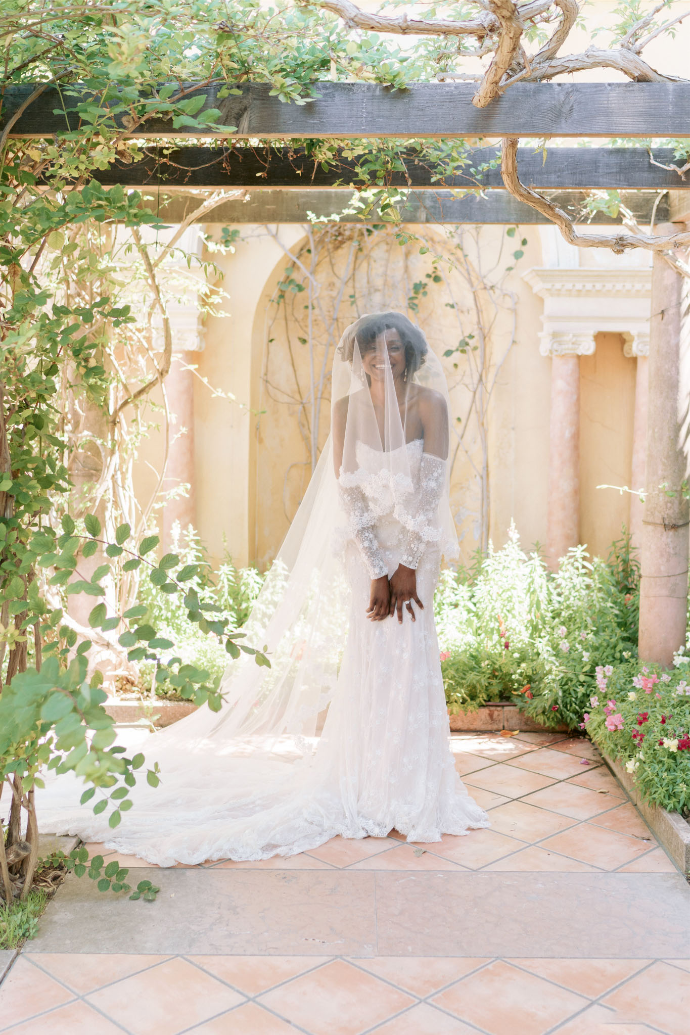 Bride in ivory lace A-line gown with cathedral veil beneath vine-covered pergola at Mediterranean courtyard