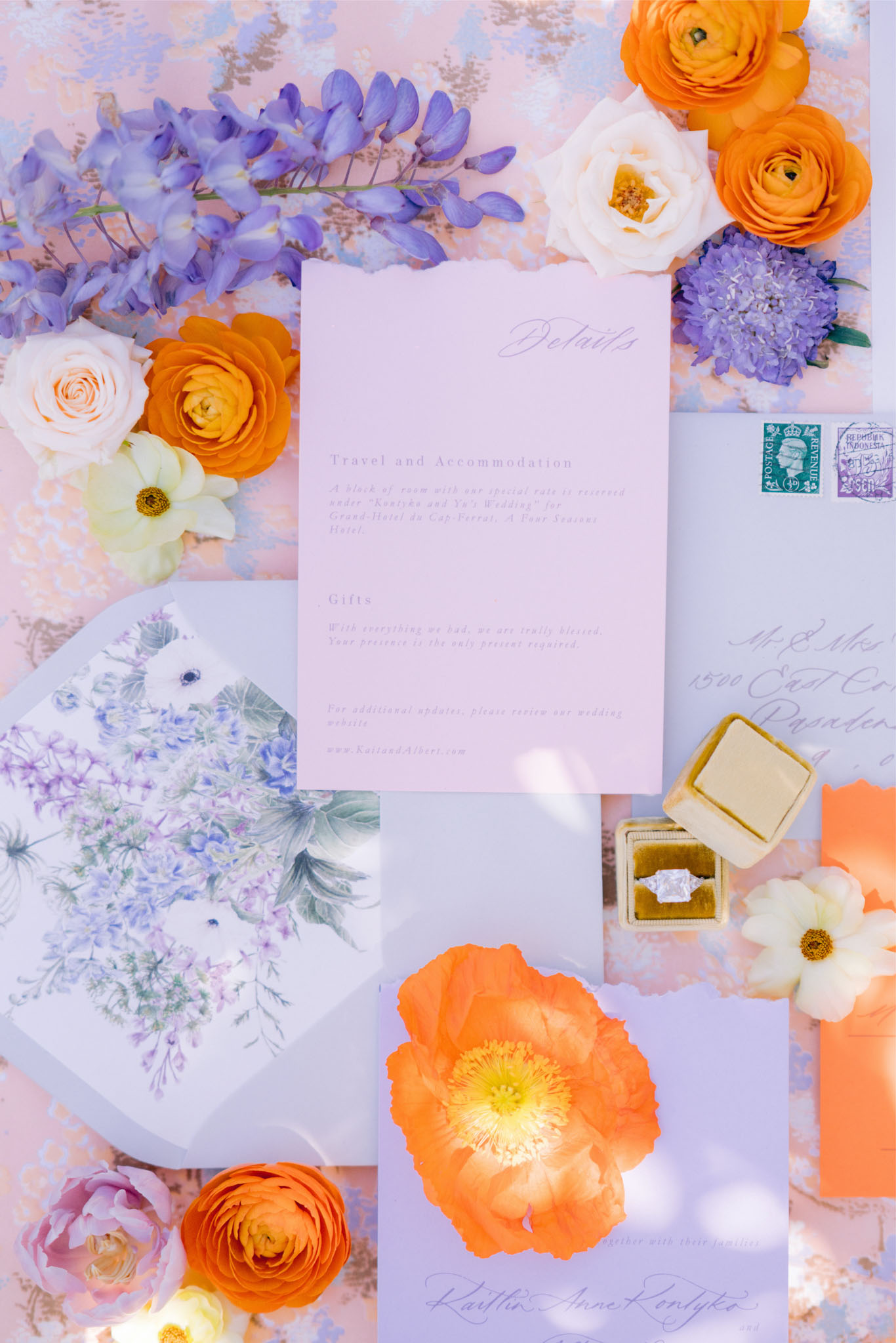 A flat lay detail shot of a wedding stationery suite arranged with fresh florals and accessories. The stationery set includes a lavender details card with script and printed text covering travel, accommodation, and gifts information, a periwinkle blue envelope with a watercolor floral botanical liner featuring wisteria and white anemones in blue and sage tones, an orange envelope, and a hand-addressed outer envelope with vintage postage stamps. The color palette is vibrant, combining lavender, periwinkle, and coral-orange across the paper goods. Scattered florals include orange ranunculus, orange Iceland poppies, cream garden roses, white anemones, purple wisteria sprigs, and purple hydrangea blooms. A mustard yellow velvet ring box holds a large cushion-cut diamond engagement ring, placed to the right of the stationery. The overall styling theme is colorful and garden-inspired with a bold complementary orange and purple palette.
