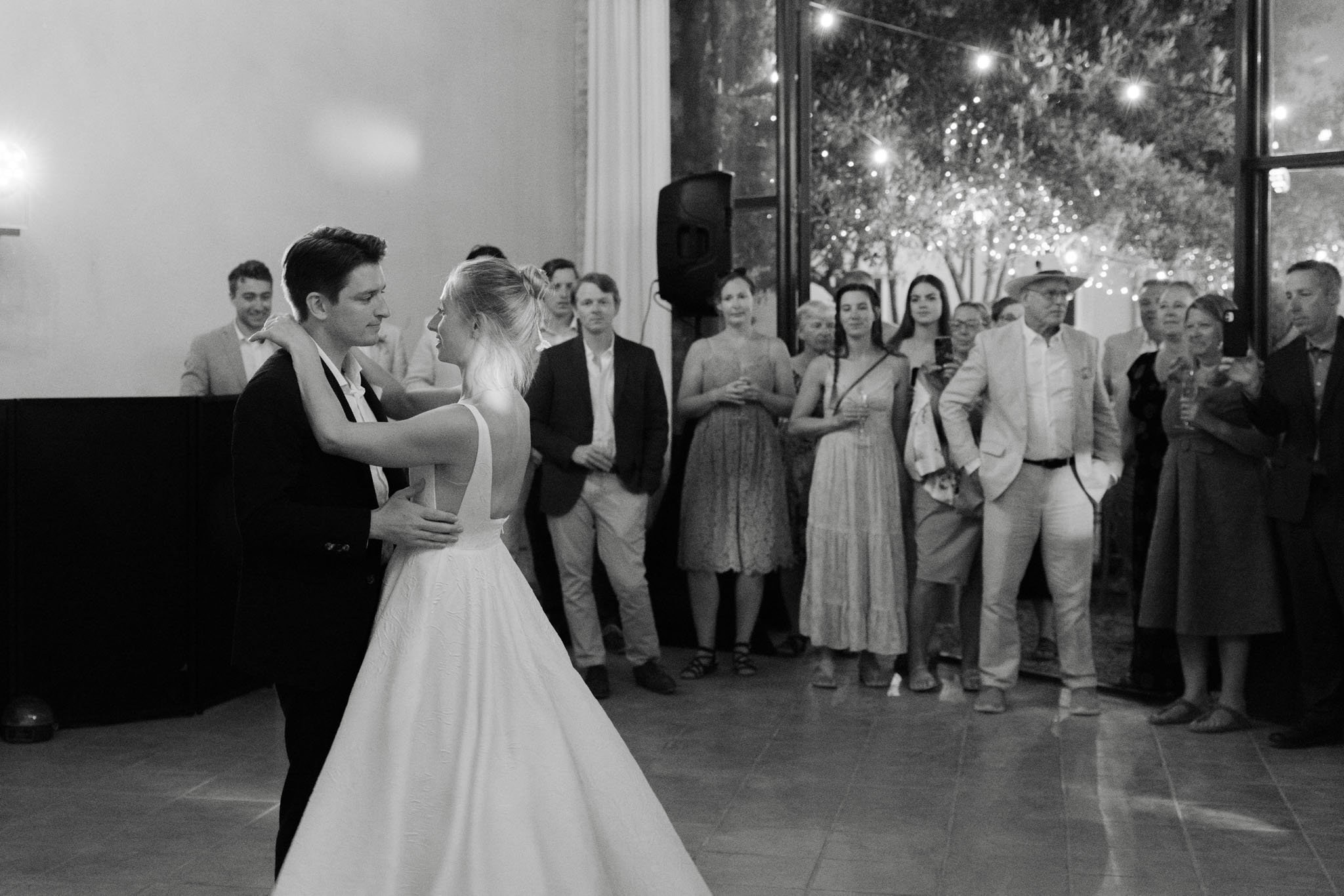 Couple shares first dance as 15 guests watch in semicircle with string-lit terrace beyond in B&W