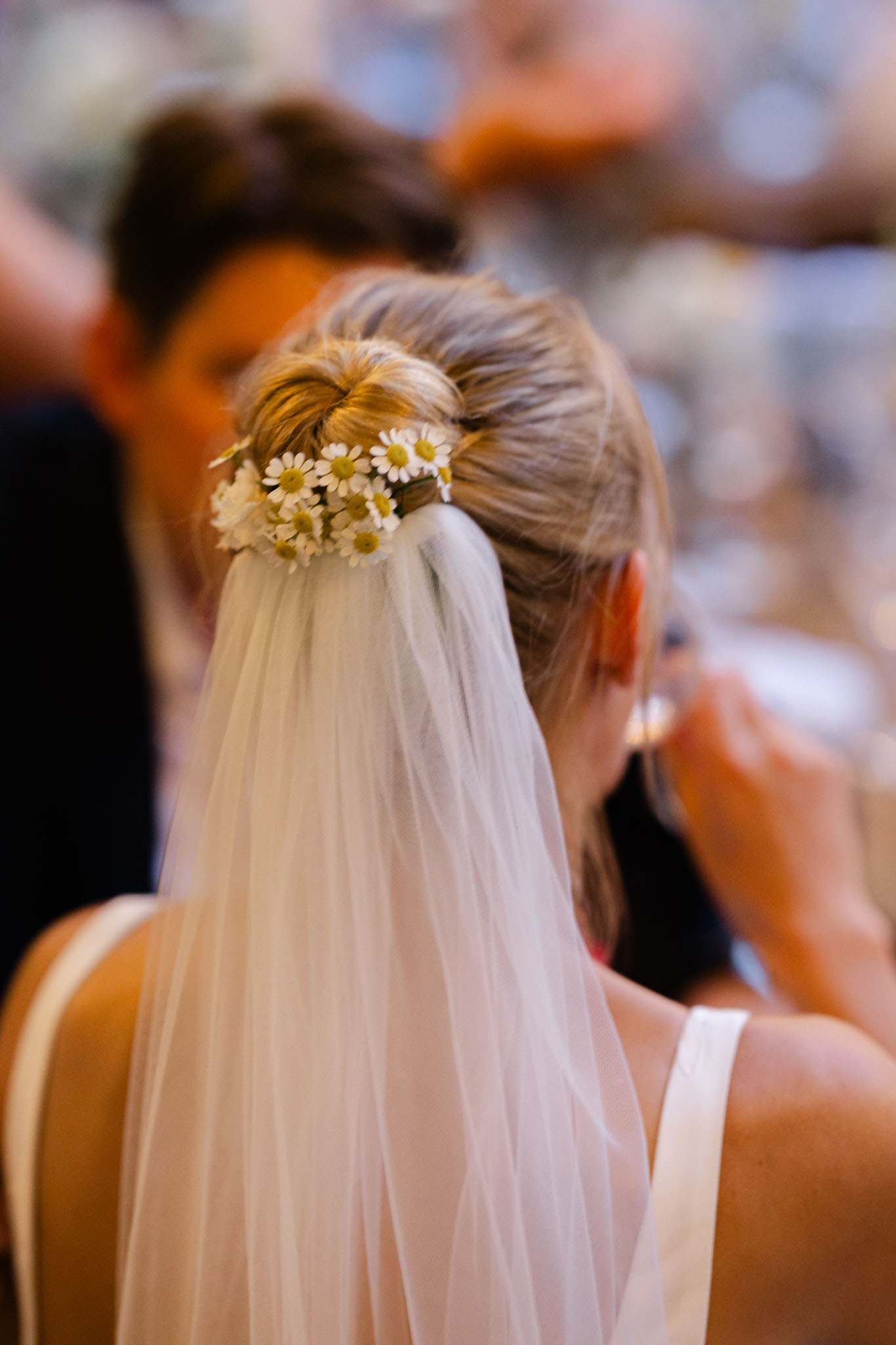 Close-up detail portrait shot of a bride's hair and veil, photographed from behind. Her blonde hair is styled in a low updo secured with a cluster of small white daisy flowers with yellow centres, and a sheer ivory veil flows from the same point. She is wearing a white sleeveless dress with a wide strap visible at the shoulder. The background is heavily blurred, showing warm tones and the suggestion of other guests, indicating this was taken during a ceremony or reception in a warm indoor or covered setting. The styling has a casual, natural feel with the daisy hair adornment as the intentional focal point.
