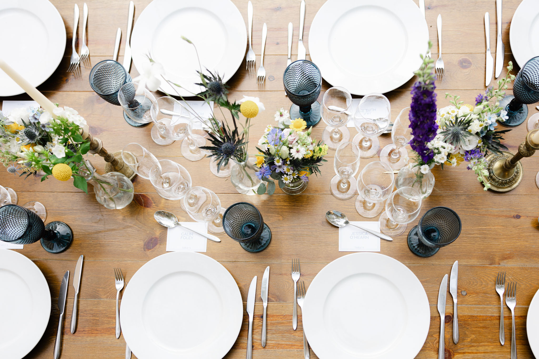 Overhead view of farm table reception setting with slate blue goblets, yellow craspedia, blue thistles, and brass candlest...