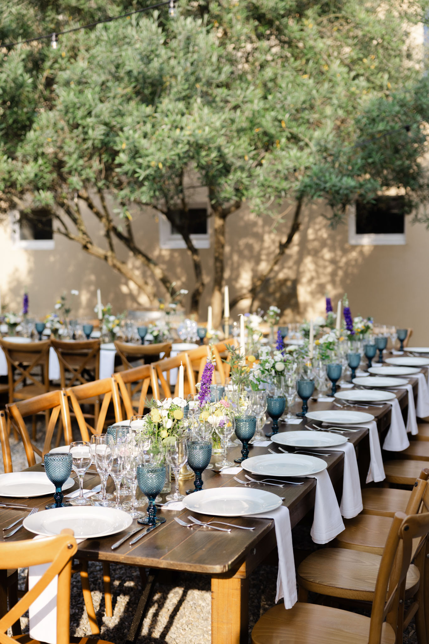 Long reception tables in courtyard with blue goblets, taper candles, and purple and white floral centerpieces