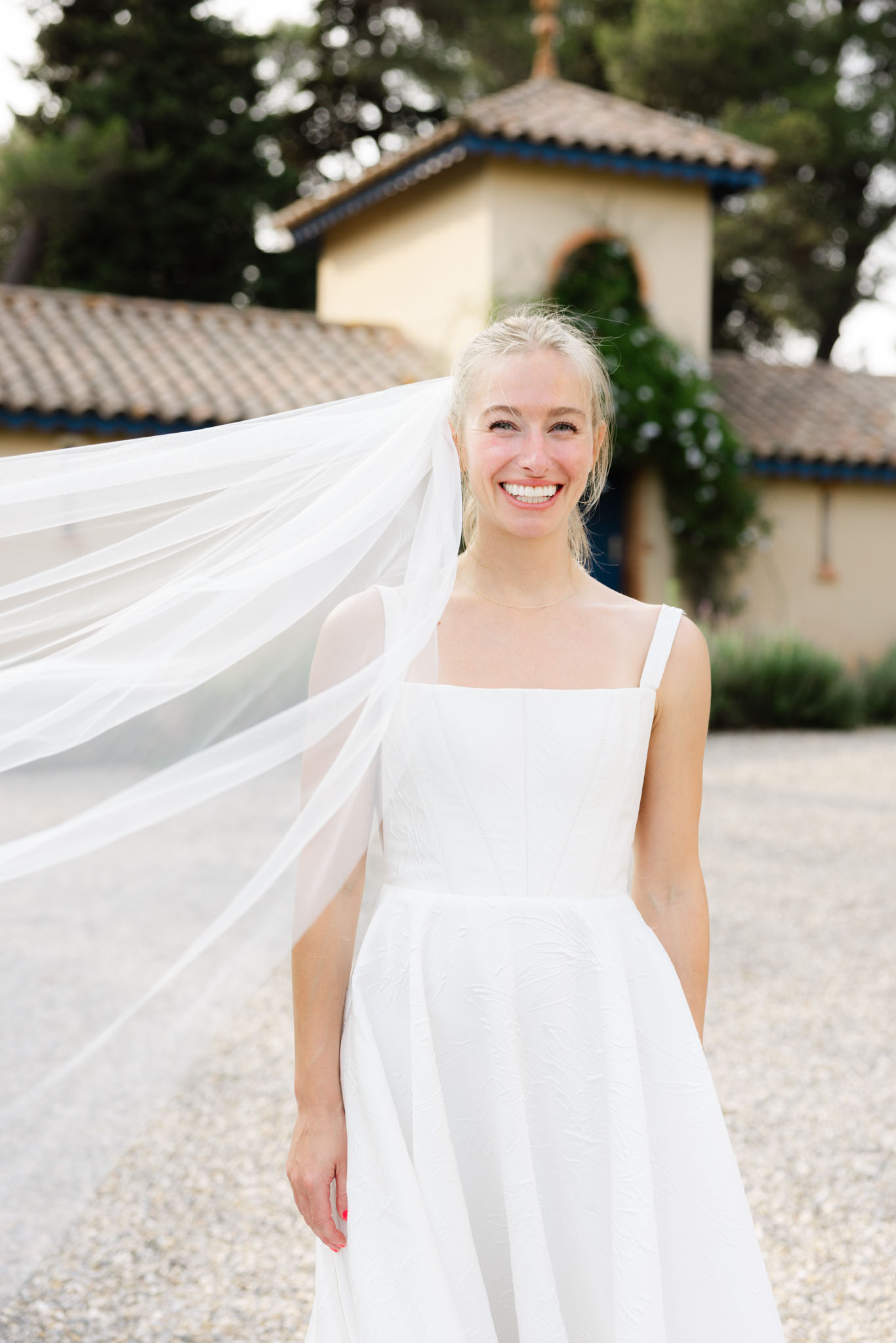 A bridal portrait taken outdoors on a gravel courtyard, featuring the bride alone in a close-to-medium shot. She wears a white structured gown with a square neckline, thick straps, and a textured jacquard-style skirt, paired with a long flowing veil caught mid-movement by the wind. Her blonde hair is loosely pulled back, and she wears a delicate chain necklace with coral-red nail polish. The background shows a Provençal-style building with ochre-yellow rendered walls, terracotta roof tiles with blue trim detailing, and an arched doorway, consistent with a French domaine or château property. The overall styling is clean and modern with a minimal aesthetic.