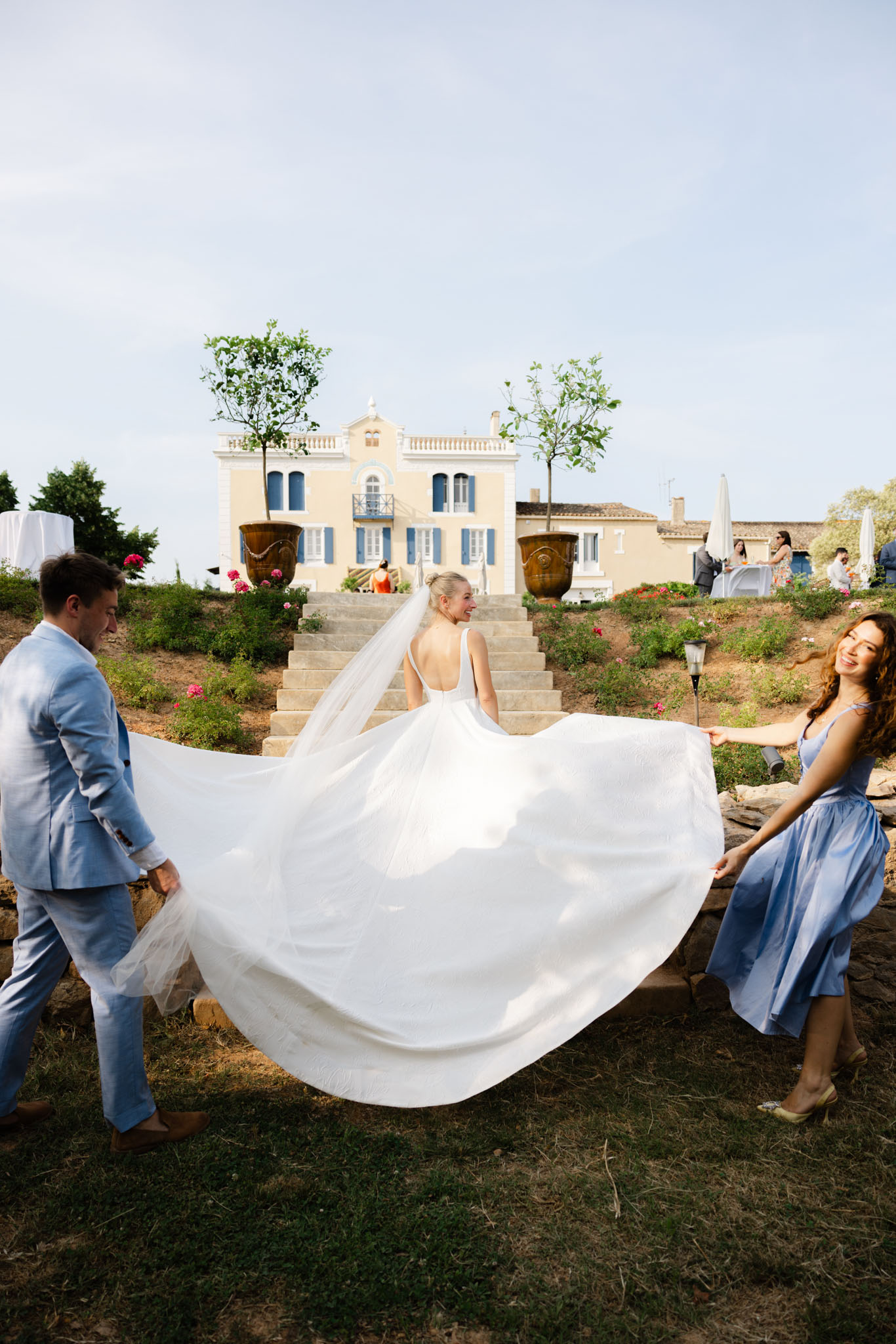 Bride ascending villa steps as attendants in blue hold billowing ballgown skirt aloft