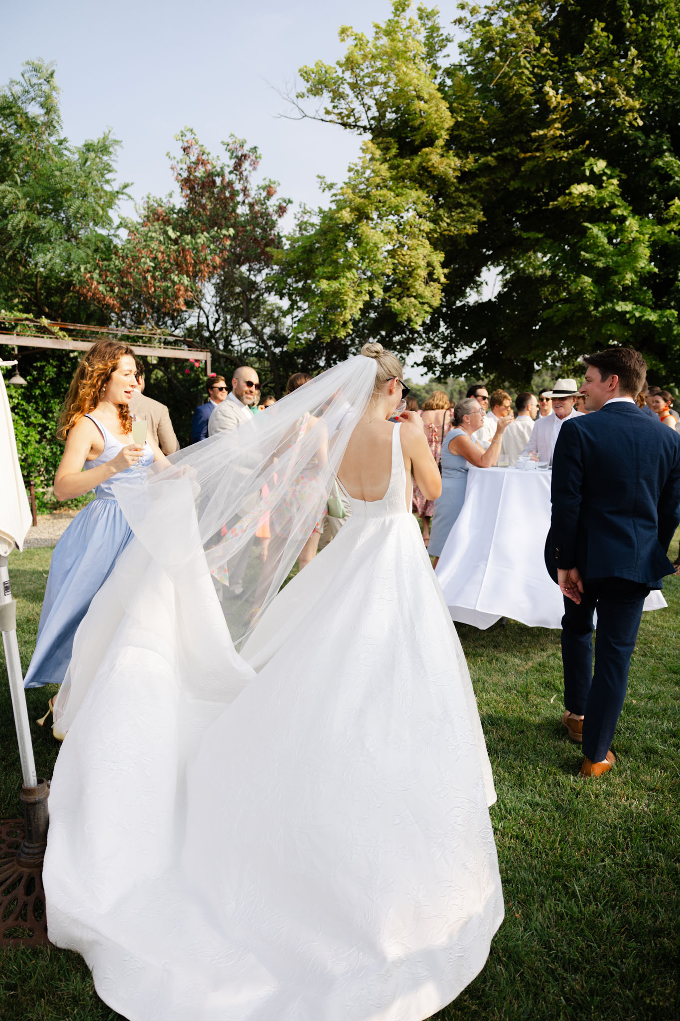 Bride with cathedral veil mingling with guests on lawn during cocktail hour, bridesmaid in blue holding veil
