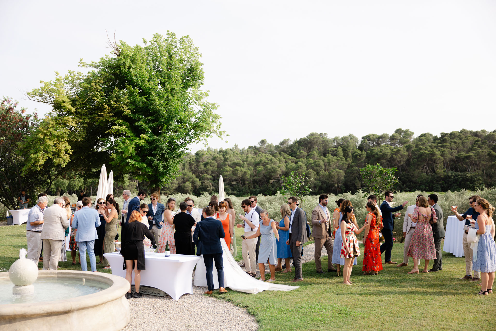 Forty-plus guests mingle on Provencal lawn with bride at cocktail table near stone fountain
