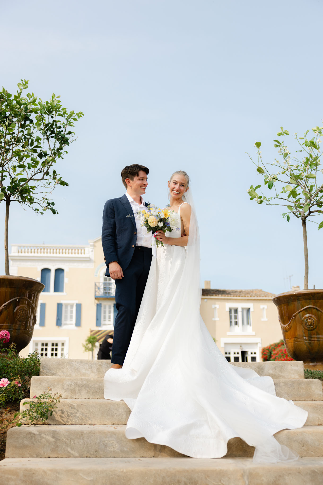 Couple on stone steps before yellow ochre manor with bride holding yellow dahlia and blue wildflower bouquet