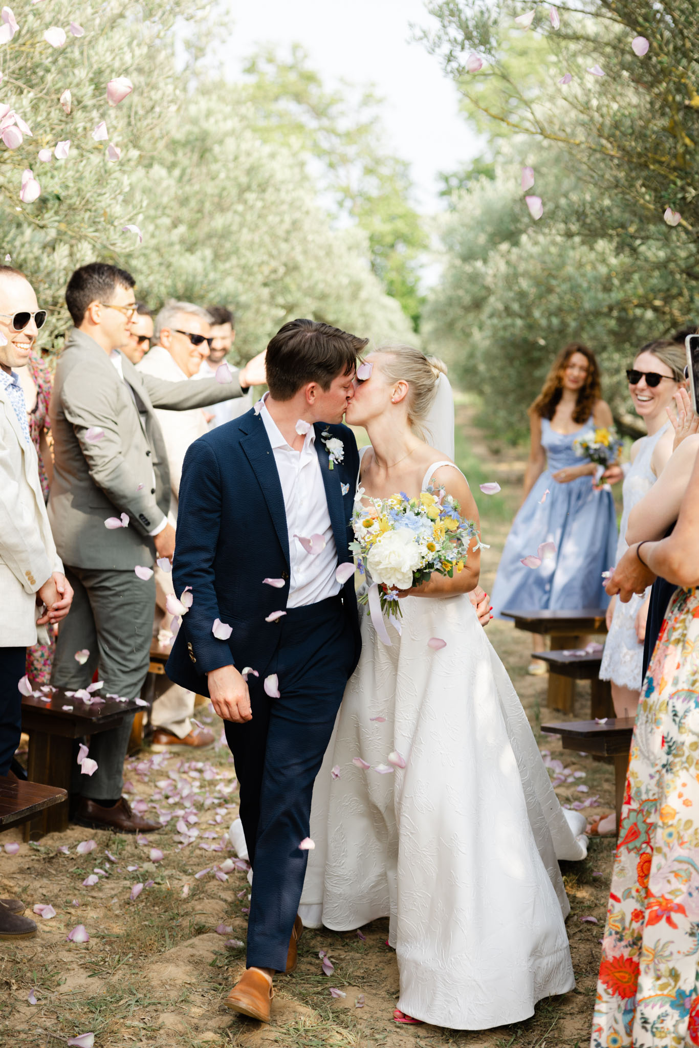 The bride and groom share a kiss while walking back up the aisle after an outdoor ceremony held in an olive grove, with approximately 15–20 guests lining either side. Pink rose petals are being tossed in the air and are scattered across the dirt aisle floor. The groom wears a navy suit with a white shirt and tan leather shoes, with a small white boutonnière. The bride wears a white textured jacquard ball gown with a veil and carries a loose bouquet of white peonies, yellow chamomile-style blooms, and blue delphinium. A bridesmaid in a powder blue midi dress is visible behind the couple, holding a matching bouquet. Guests are dressed in a mix of casual summer attire including floral prints and linen suits. The setting is rustic and natural, with wooden bench seating along the aisle. Wide shot, mid-length perspective capturing both the couple and the surrounding guests.
