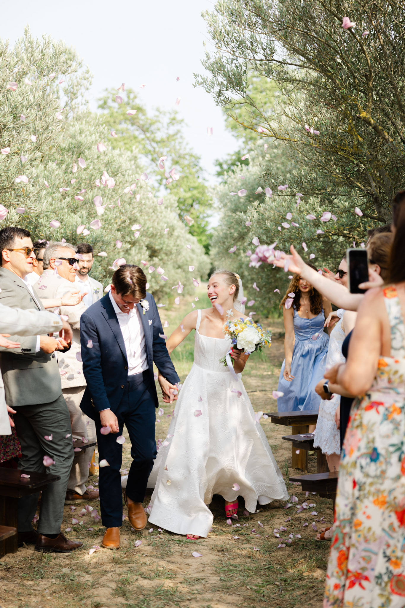 The couple is walking back down the aisle during their outdoor ceremony recessional, surrounded by guests tossing light pink and lavender rose petals into the air. The ceremony took place in an olive grove, with rows of wooden benches lining a sandy dirt aisle flanked by olive trees. The bride wears a white square-neck A-line gown with textured fabric and carries a bouquet of white peonies, yellow and blue wildflowers, and greenery; she is laughing joyfully. The groom wears a navy suit with brown leather shoes and a small boutonniere. Guests on both sides are dressed in a mix of summer outfits including a powder blue fit-and-flare dress, a sage green suit, a floral print dress, and linen separates. The composition is a medium wide shot taken from ground level, capturing the couple at center with guests blurred on either side and petals suspended mid-air.