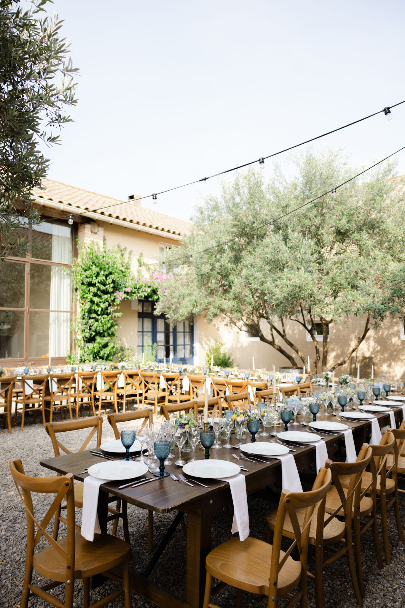 An outdoor wedding reception setup in a courtyard of a French mas-style property with warm ochre rendered walls and blue-framed windows. Multiple long farm-style tables in dark wood are arranged in rows on a gravel surface, surrounded by natural wood cross-back chairs. The tables are dressed with white linen napkins, white ceramic plates, clear stemware, teal-blue hobnail goblets, tall white taper candles in brass holders, and low floral clusters featuring small yellow, purple, and white wildflower-style blooms. Festoon string lights are strung overhead across the courtyard. The overall styling palette combines warm wood tones, white, and dusty teal with a relaxed Provençal aesthetic. Wide shot capturing the full reception layout. Potential venue feature image.