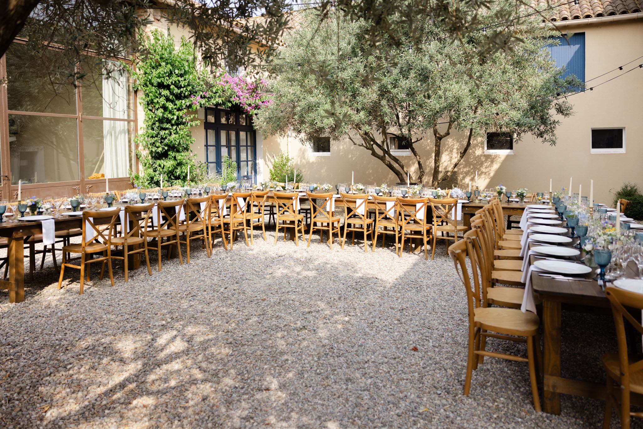 An outdoor wedding reception setup photographed in a gravel courtyard enclosed by warm beige rendered buildings with blue-painted window frames and doors. Three long wooden farmhouse-style tables are arranged in a U-shape configuration, seated with natural oak cross-back chairs and white linen napkins. The tables are set with white plates, blue tinted glassware, tall white taper candles, and low floral centerpieces featuring small white and yellow blooms. Fairy lights are strung overhead, and the setting has a relaxed Provençal rustic aesthetic. Wide shot capturing the full table layout before guests arrive. Potential venue feature image.