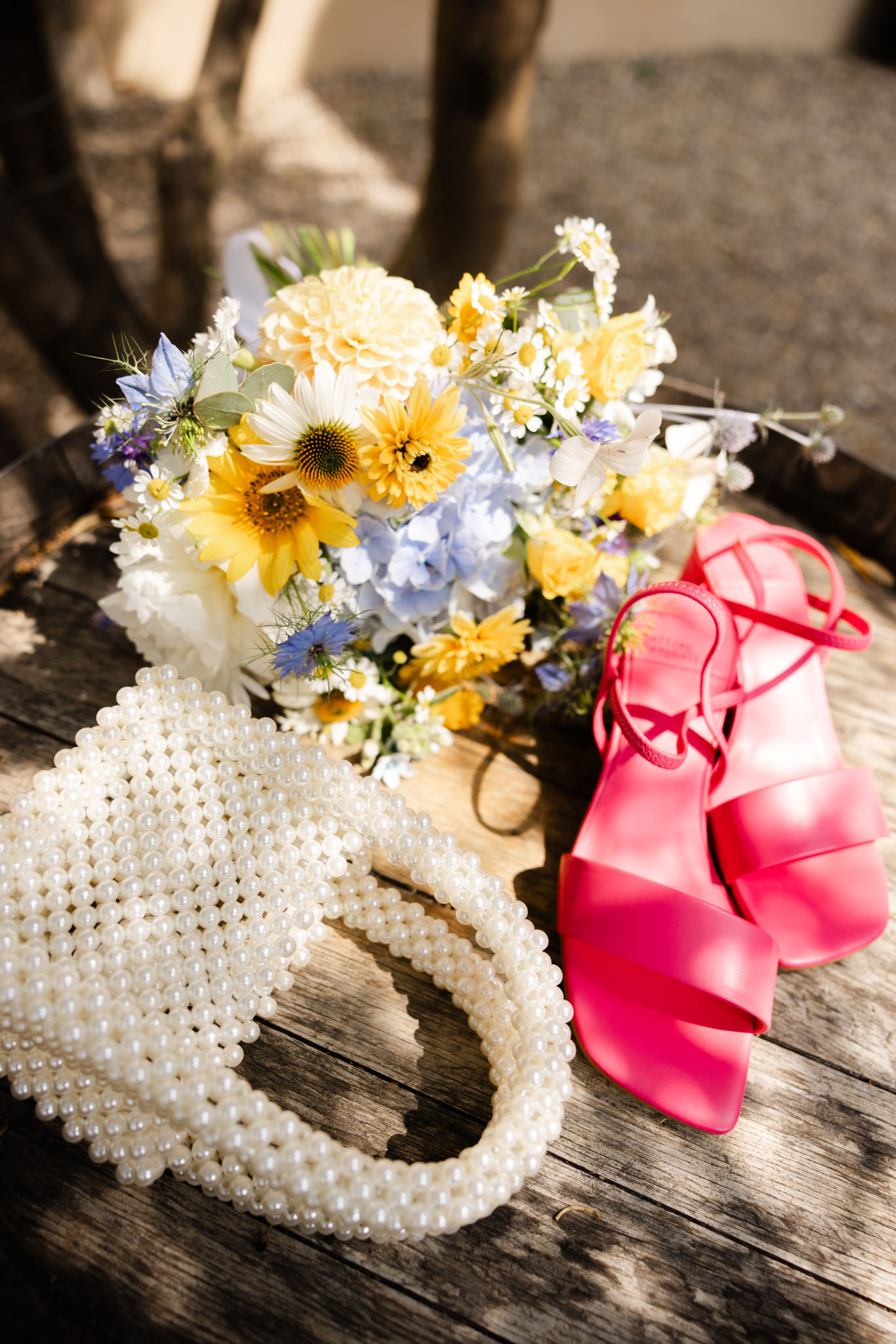 Bridal flat lay with sunflower and blue hydrangea bouquet, pearl beaded bag, and pink sandals on wooden surface
