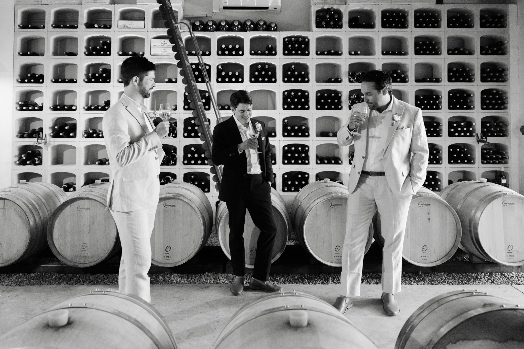 A black-and-white wide shot of three men in wedding attire tasting wine inside a winery chai or barrel room. The two men on the left and right are wearing light-colored suits, while the man in the center wears a darker suit with a floral boutonniere; all three hold wine glasses and appear relaxed and jovial. Behind them, a large white grid-style wine rack filled with bottles spans the entire wall, with a row of oak aging barrels branded with what appears to be a château name lined up in front of it. A wooden ladder leans against the wine rack in the center of the frame, and additional barrels are visible in the foreground. The image has strong contrast with bright whites and deep blacks, giving the setting a clean, graphic quality.