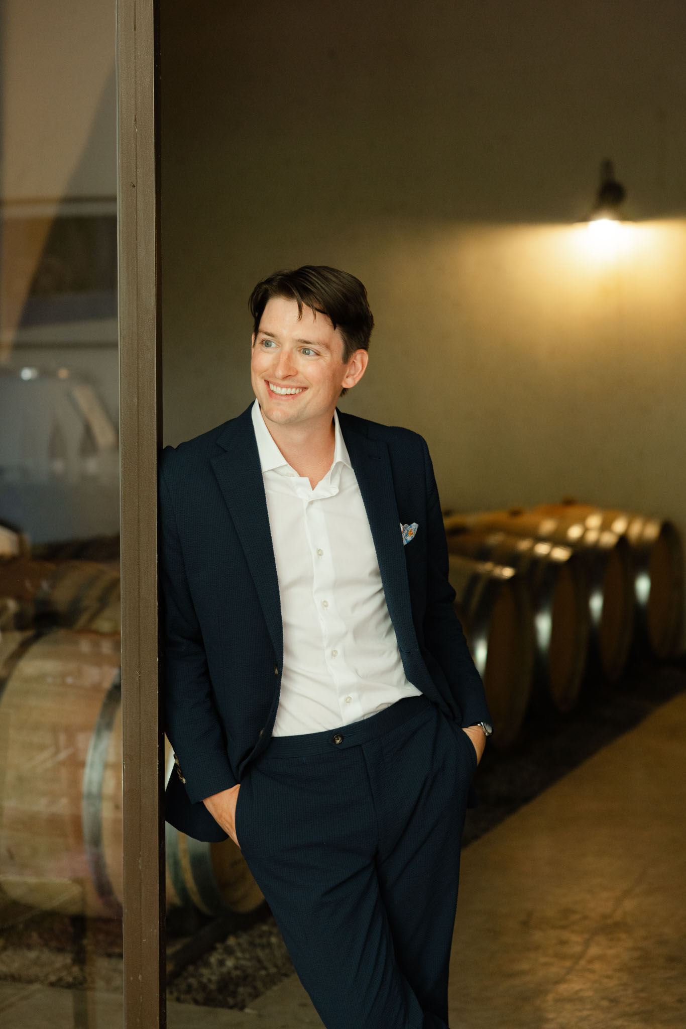 Groom in navy suit leaning against doorframe in wine barrel cellar with warm golden lighting
